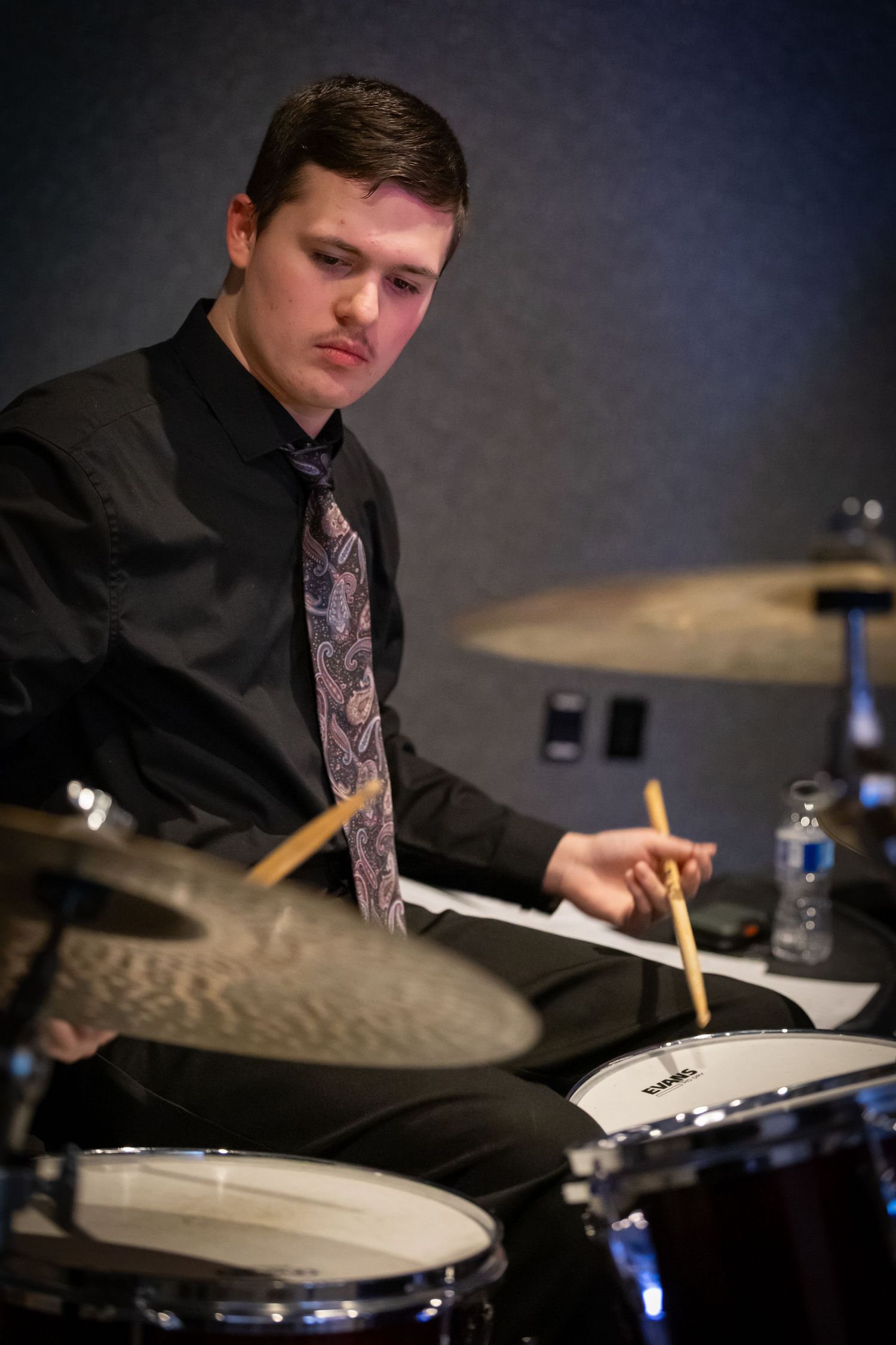 A young male drummer in a black shirt and tie playing drums with focused concentration during a jazz performance.
