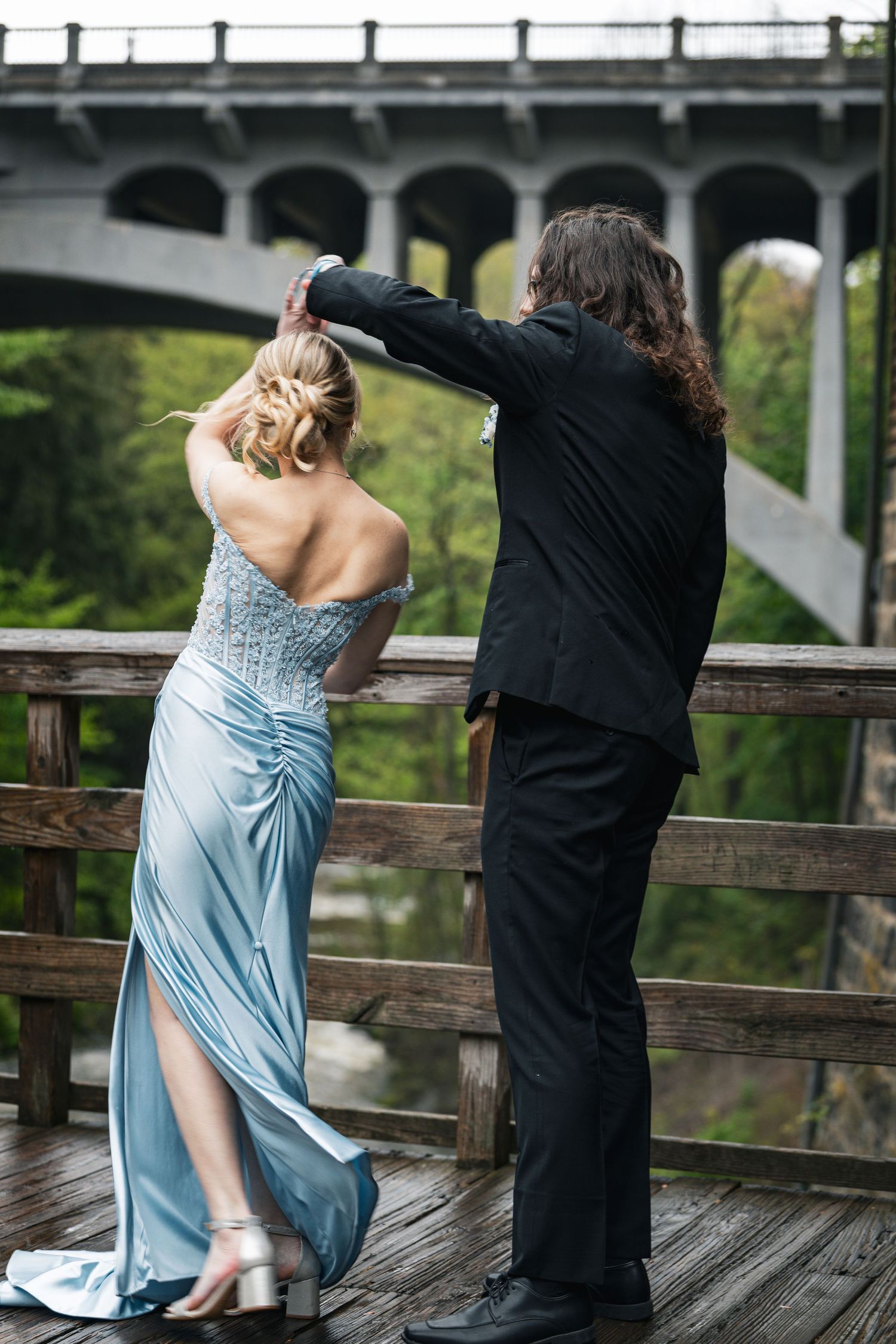 Couple in formal wear posing on wooden bridge overlook, woman in blue gown, man in black suit with long hair.