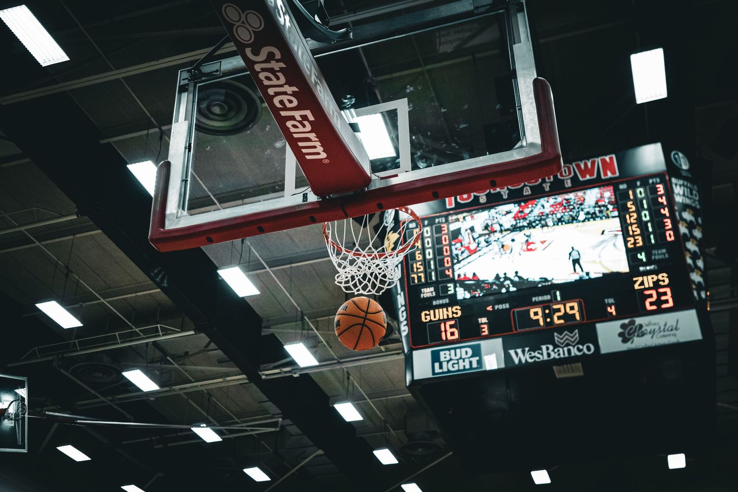 Indoor basketball court with fluorescent lighting and scoreboard visible.