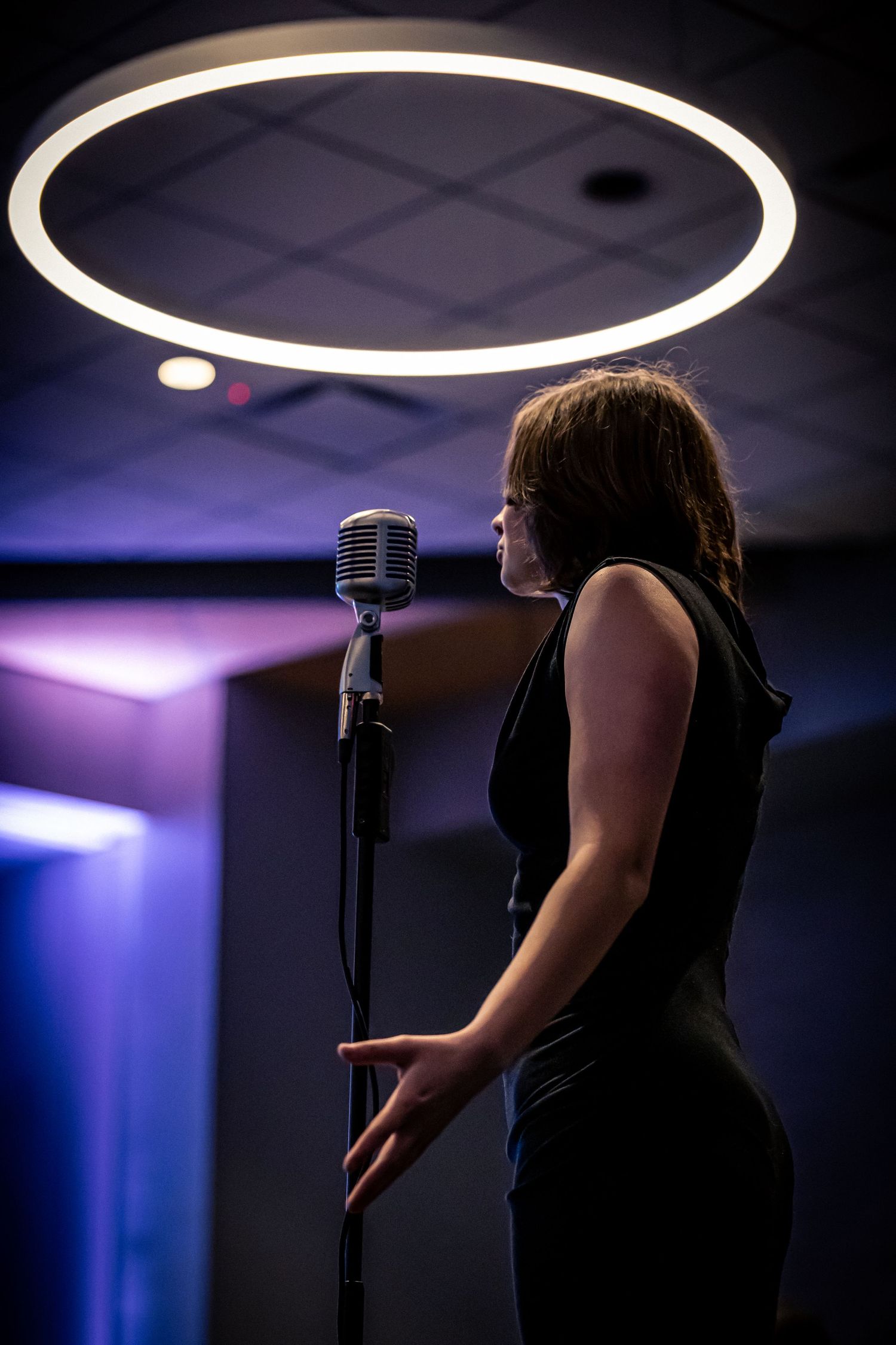 Woman in black dress performing at a retro microphone under a glowing circular light in a moody venue.