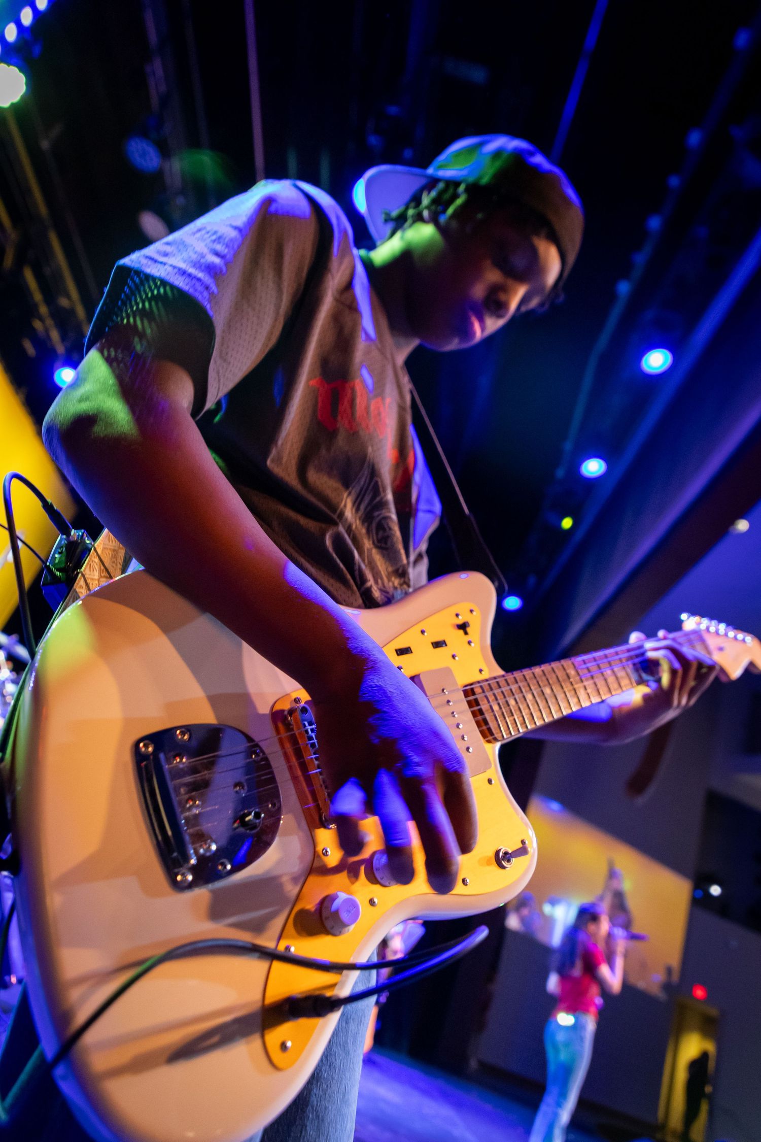 Young musician plays white electric guitar on stage under colorful blue and purple lights during live performance.