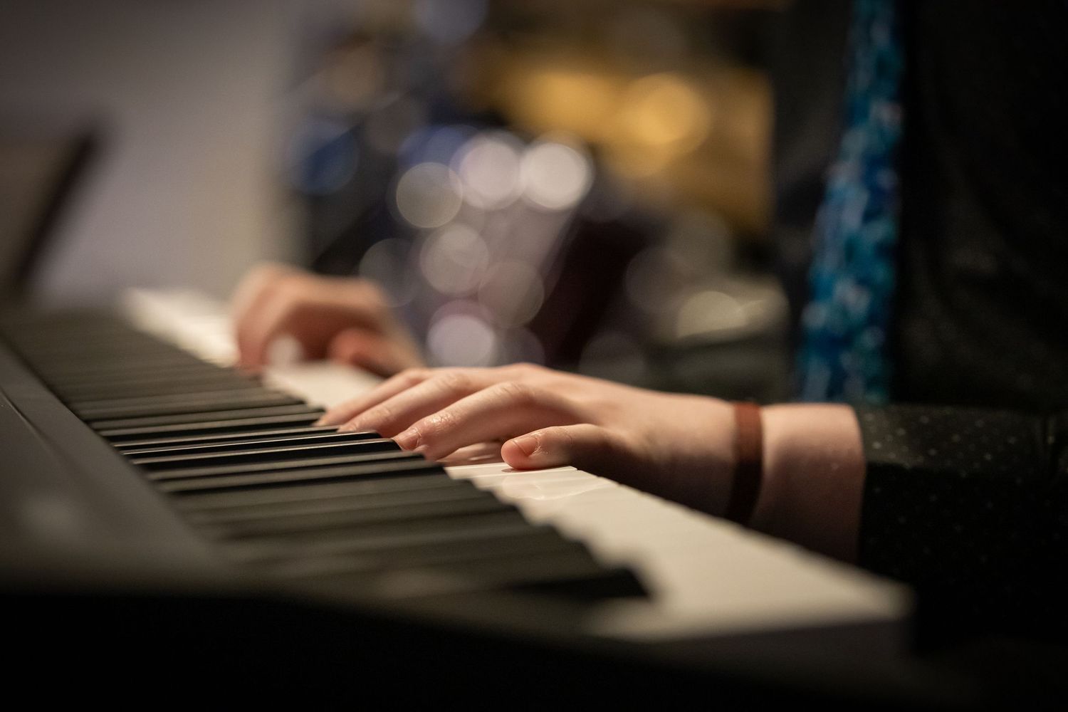 Close-up of hands playing a keyboard piano, with blurred drum kit in the background.