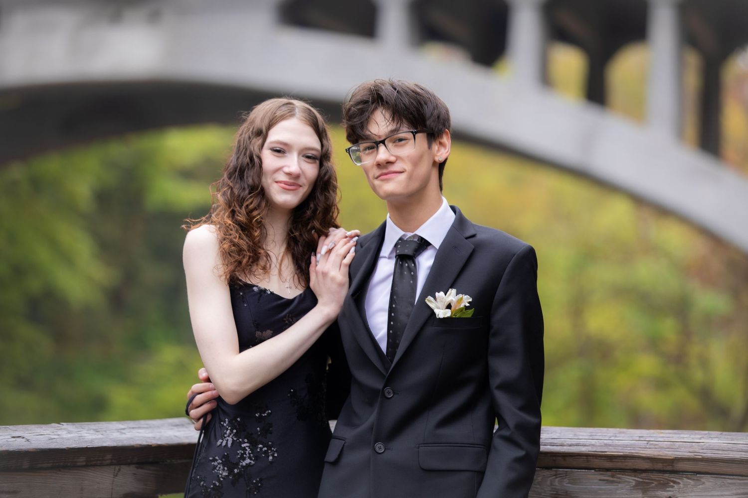 A young couple in formal prom attire poses outdoors under a stone bridge surrounded by autumn foliage.