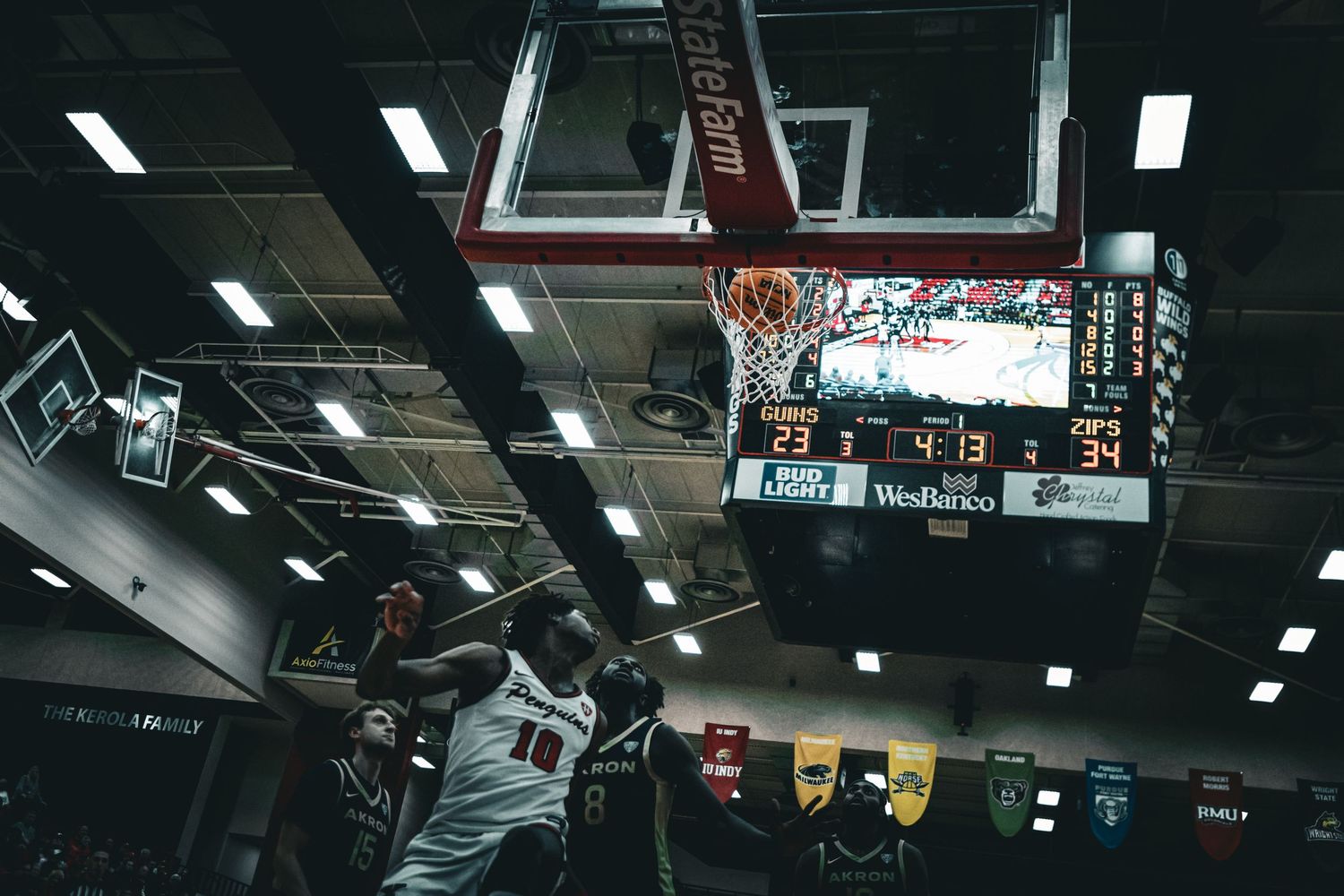 Basketball players compete in an indoor gymnasium with overhead lighting and team banners.