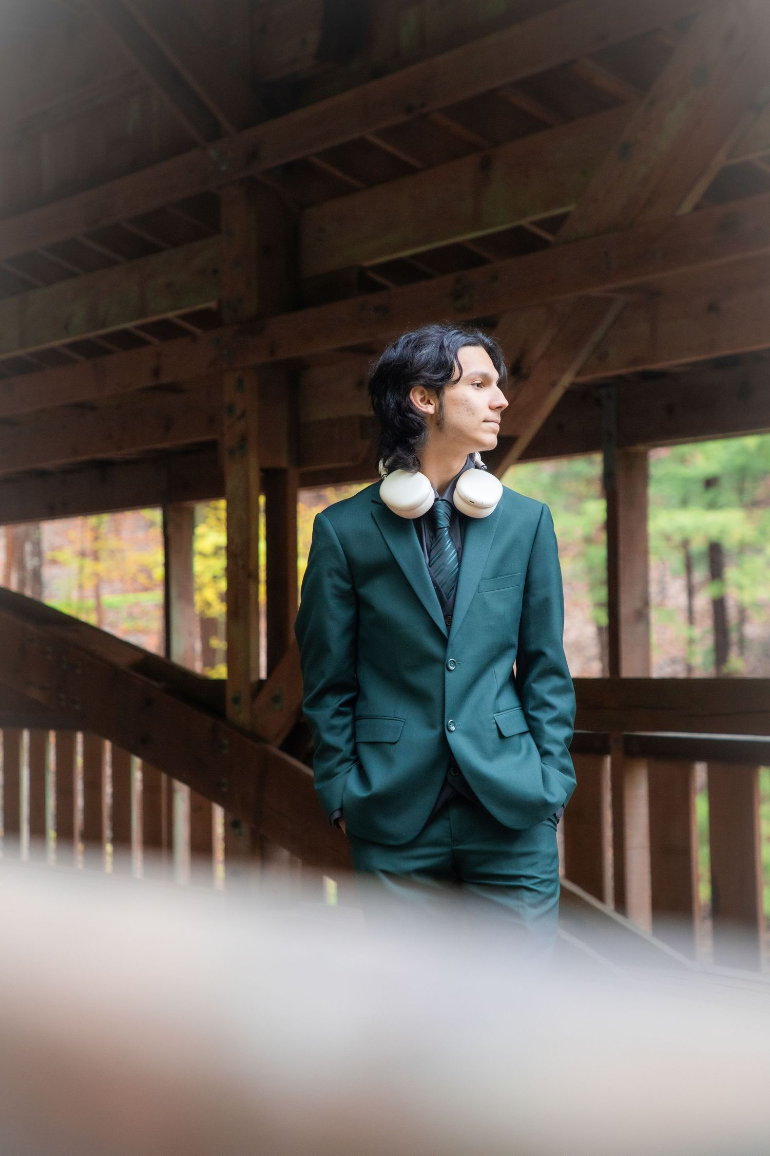 Young man in green suit with white headphones around neck standing on wooden covered bridge surrounded by autumn trees.