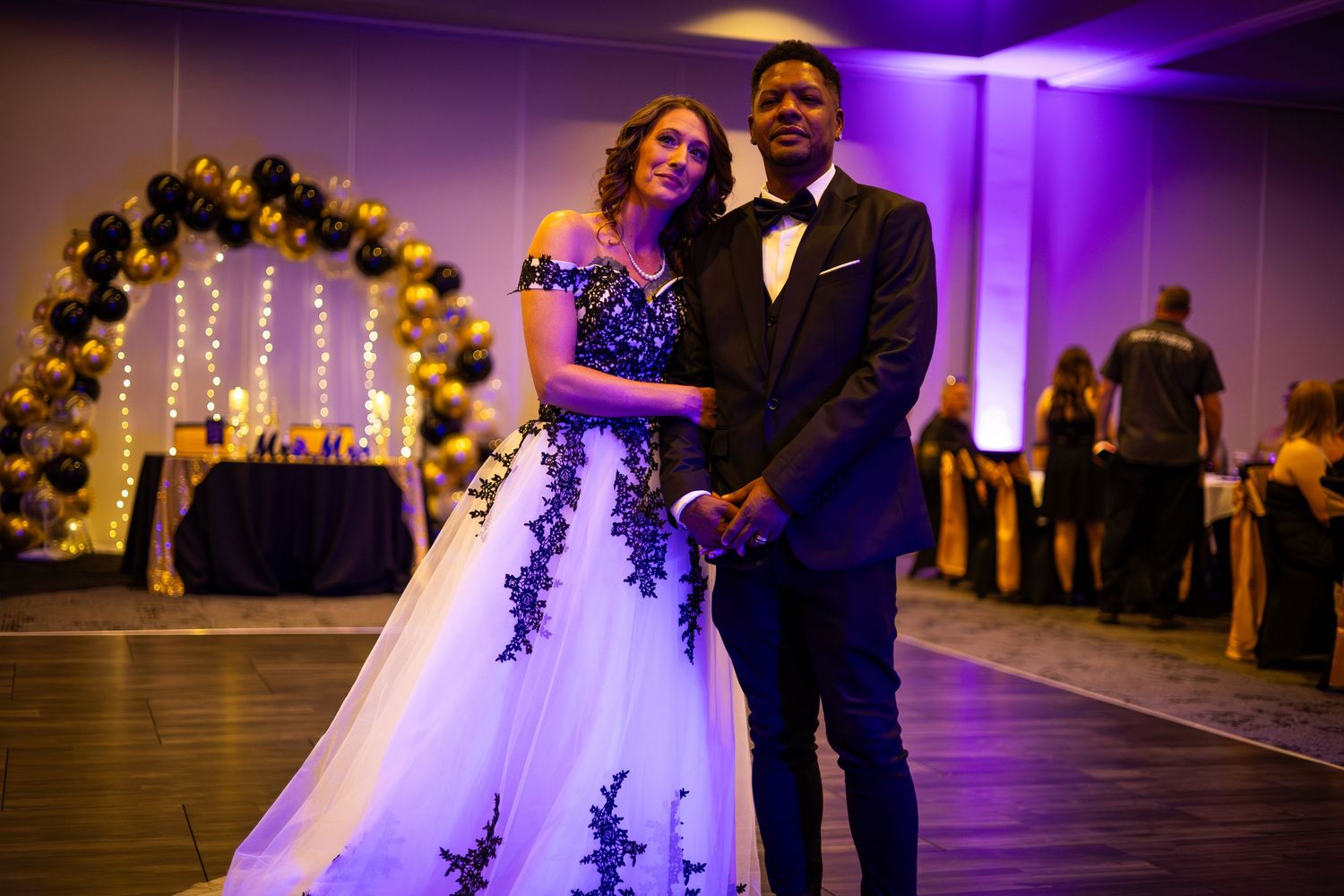 Wedding couple posing under decorative floral arch with purple lighting.