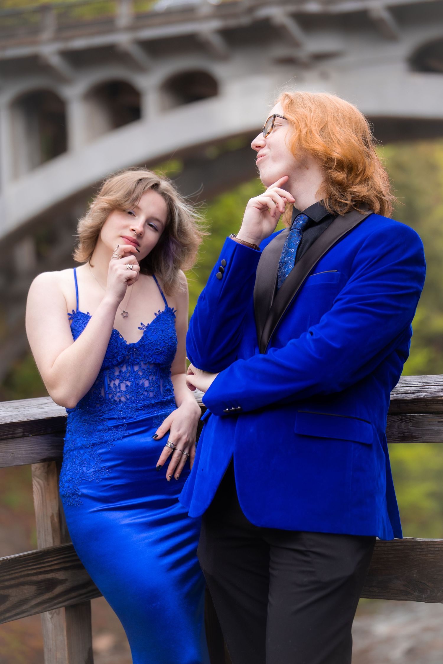 Two teens in matching blue formal wear pose thoughtfully on a wooden bridge for prom photos.