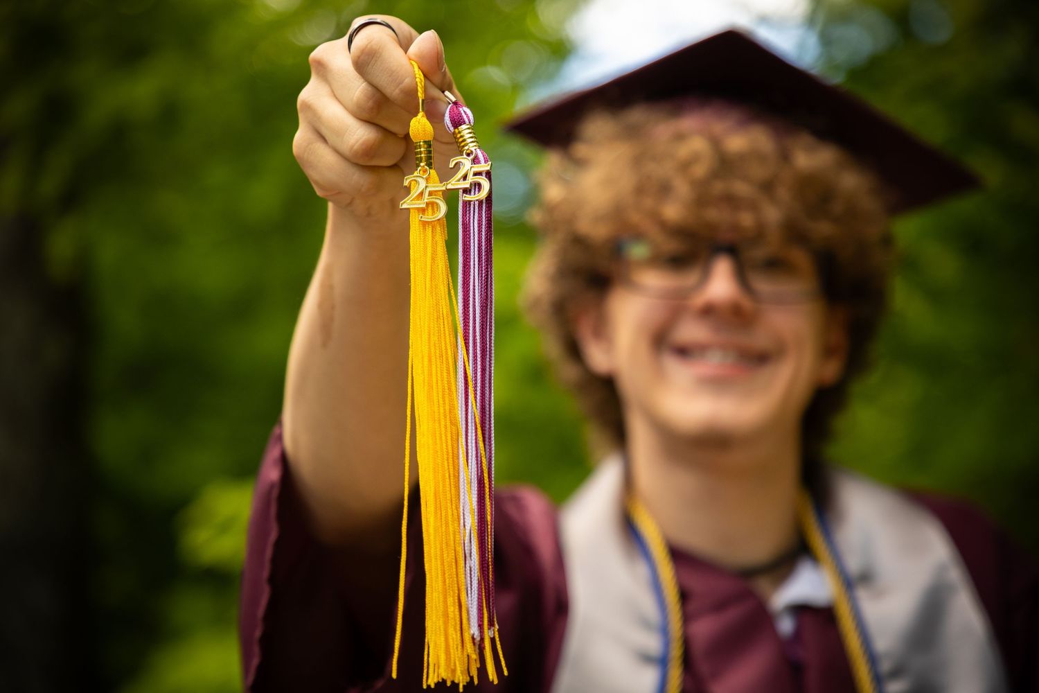 Graduate holds yellow tassel from graduation cap in outdoor setting.