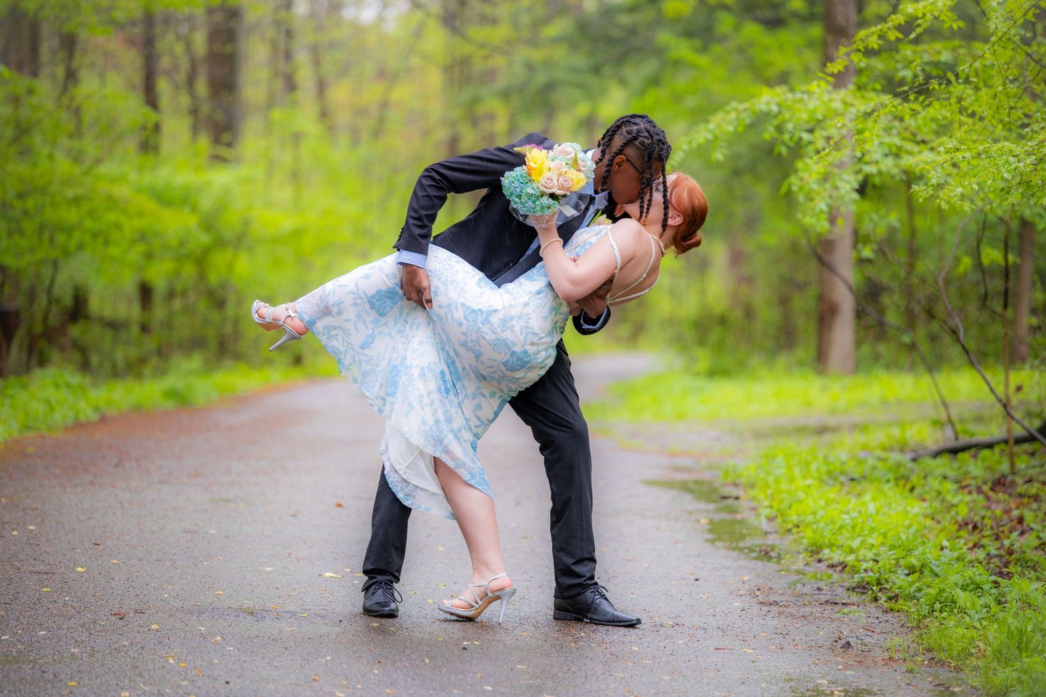 Groom dipping bride in a romantic dance on a rainy forest path, bride holding a colorful bouquet.