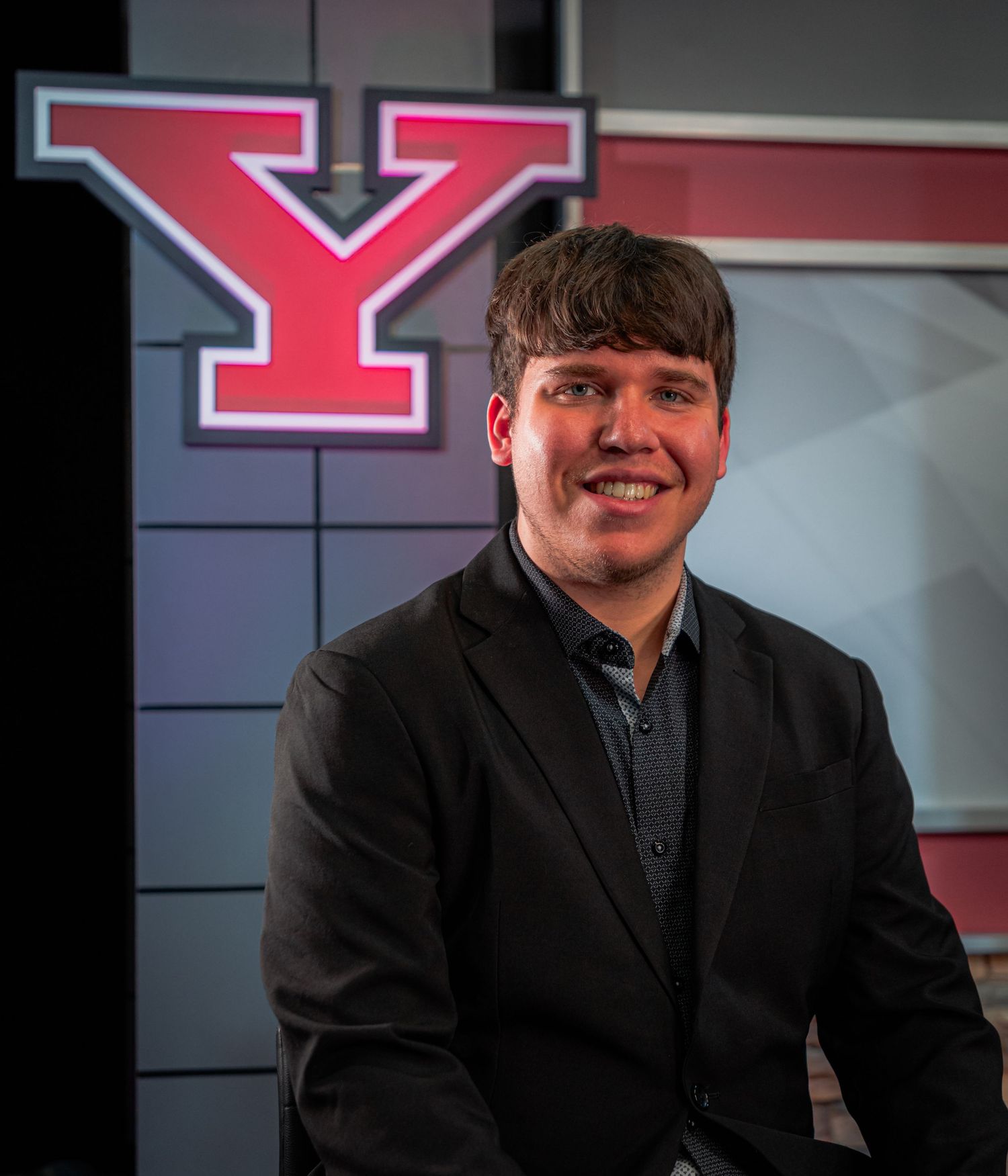 Young man in black blazer smiling in front of a red letter Y logo backdrop in a professional studio setting.