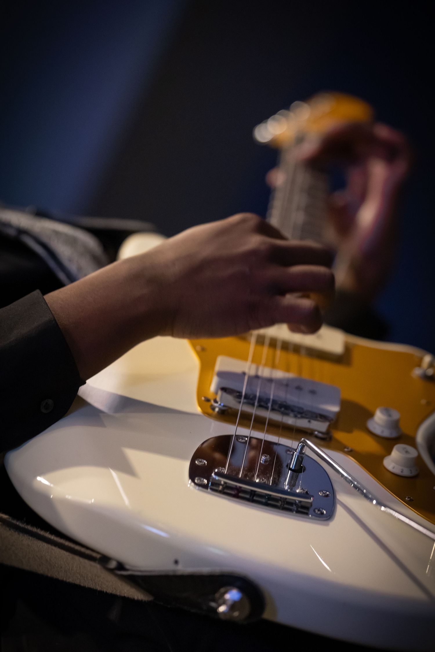 Close-up of hands playing a white and yellow electric guitar with chrome bridge in moody studio lighting.