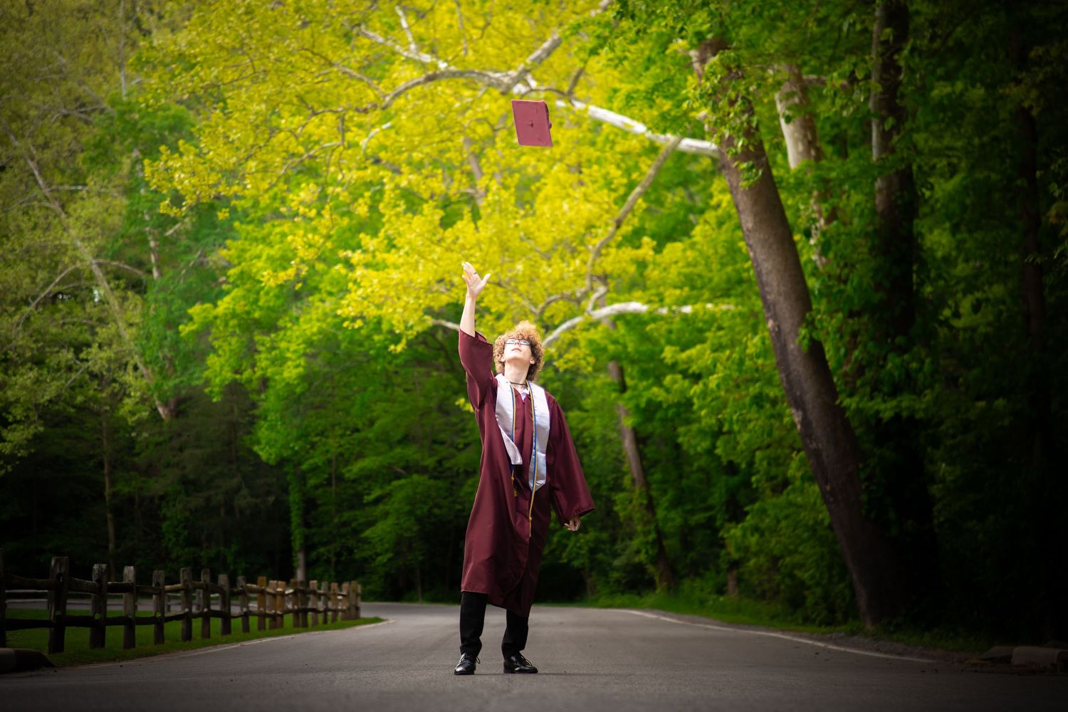 Graduate in maroon regalia celebrates by tossing diploma in air on tree-lined path.