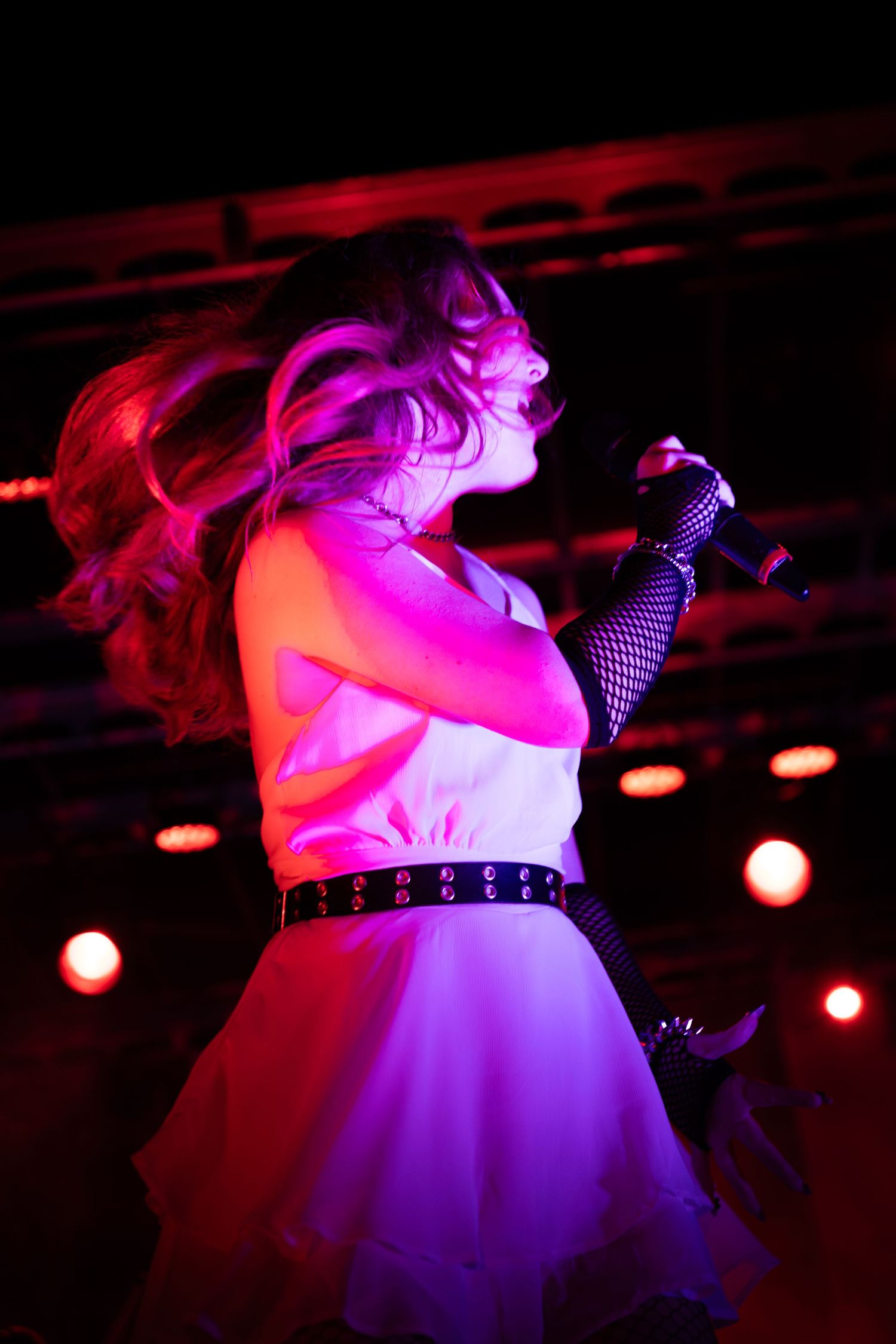 Female singer performing on stage under pink and purple lights, wearing white dress with fishnet gloves, hair flowing.