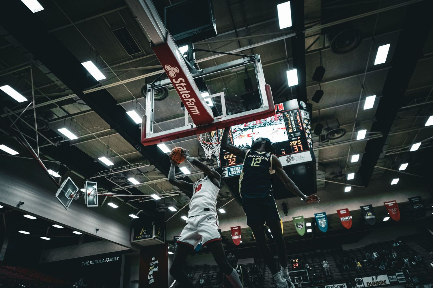Dark indoor basketball court with overhead lighting and team banners displayed on walls.