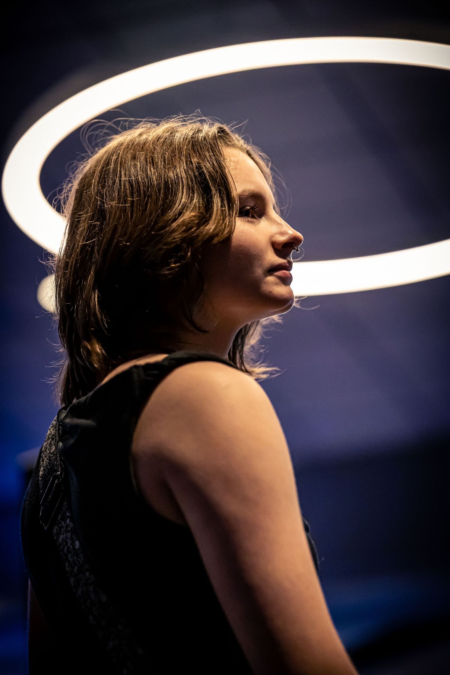 Young woman in black sleeveless top posing under a circular neon halo light in a dark blue setting.