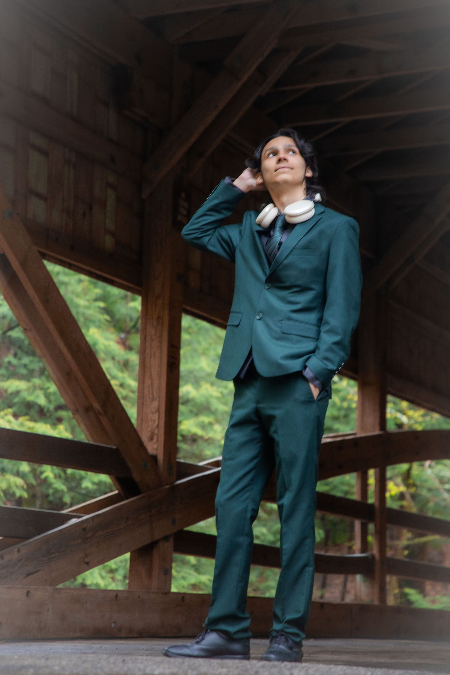 Young man in teal green suit with headphones poses thoughtfully under wooden covered bridge structure.