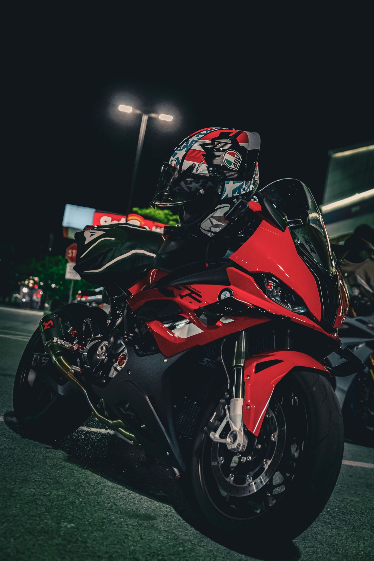 A rider in a decorated helmet sits on a red sport motorcycle under bright street lights at night.