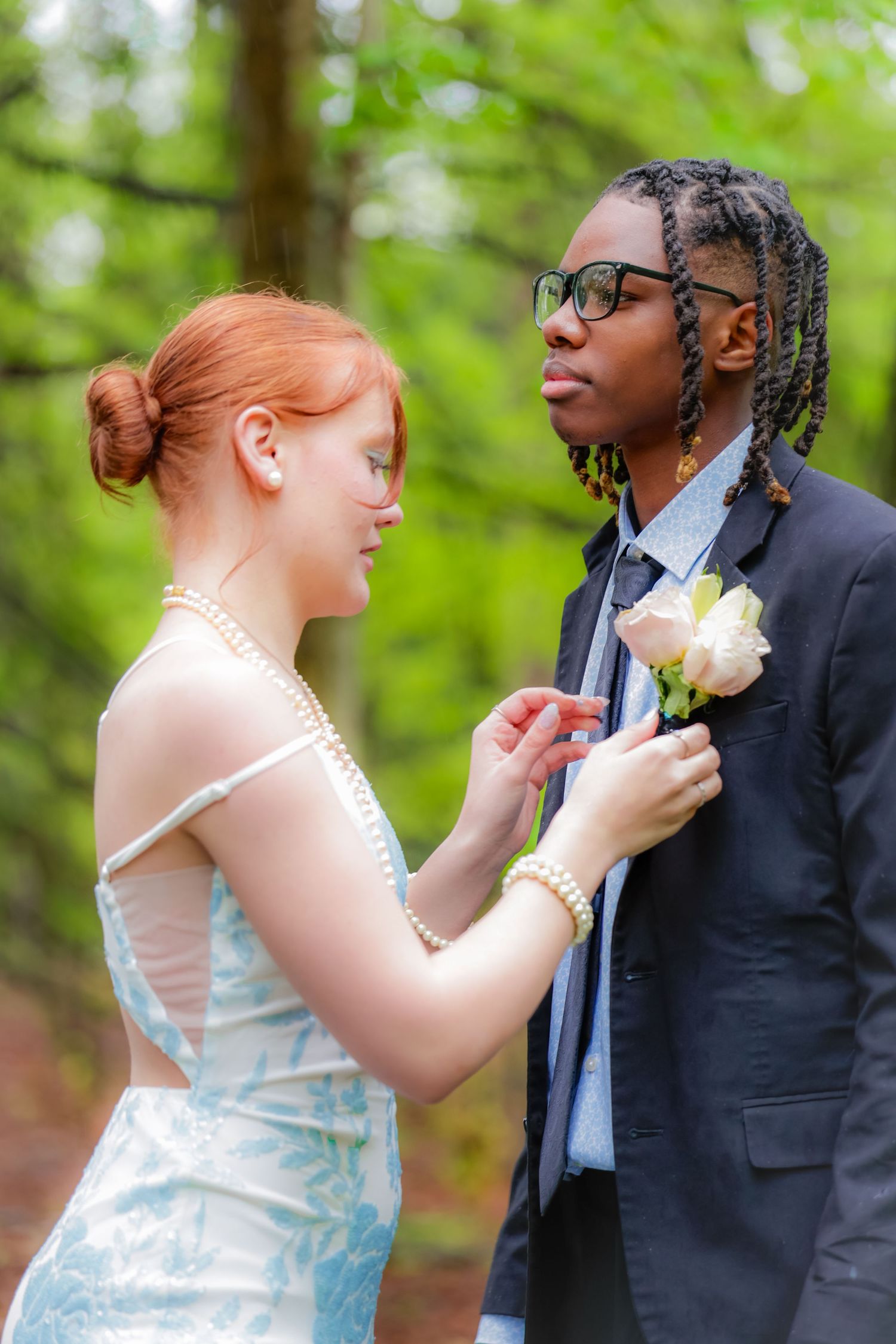 Young interracial couple in formal wear pinning a boutonniere outdoors in a green forest setting.