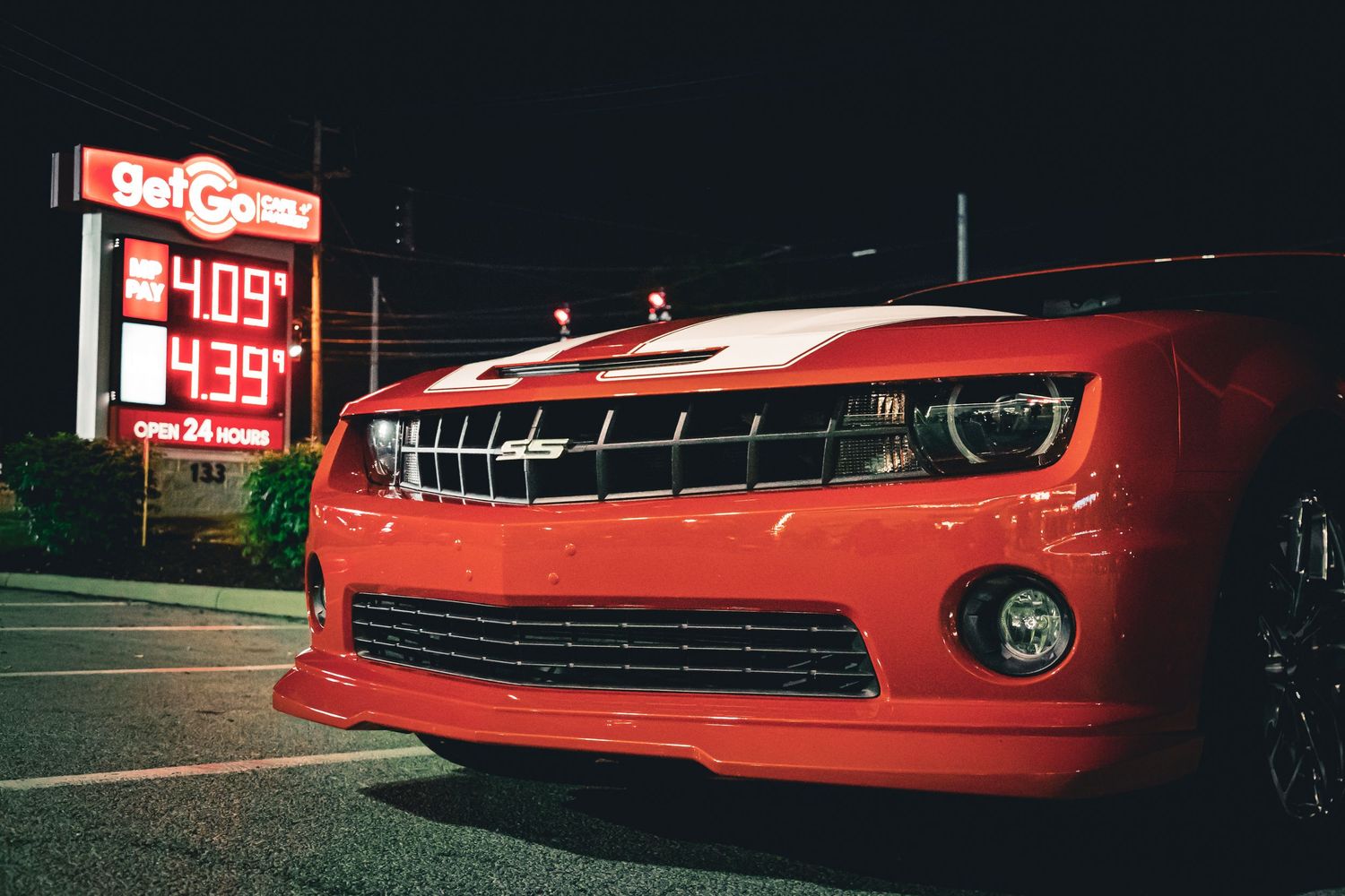 Red Chevrolet Camaro SS parked at a gas station at night with fuel prices displayed on illuminated sign.