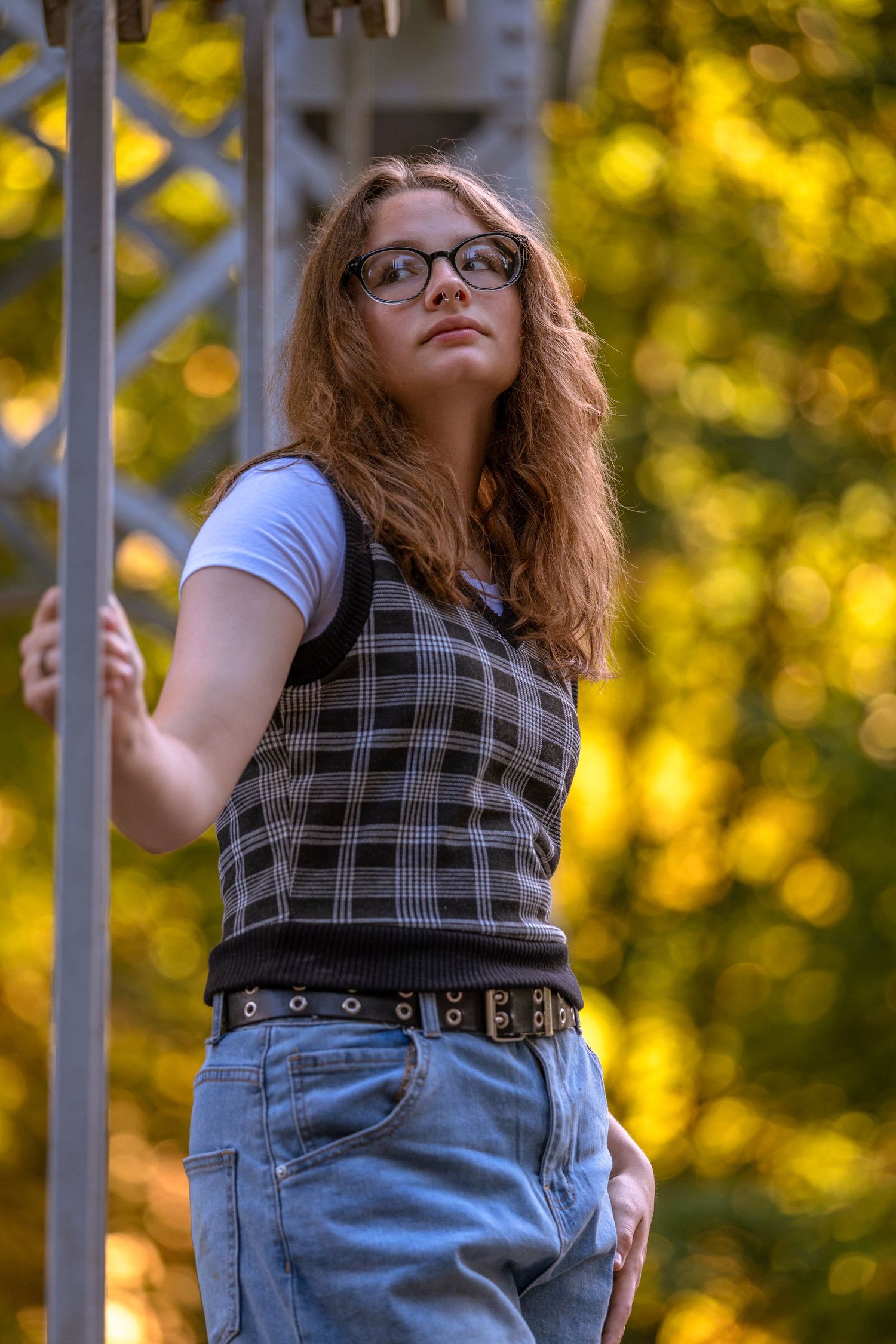 Person in plaid dress and denim poses on white railing against golden bokeh background.