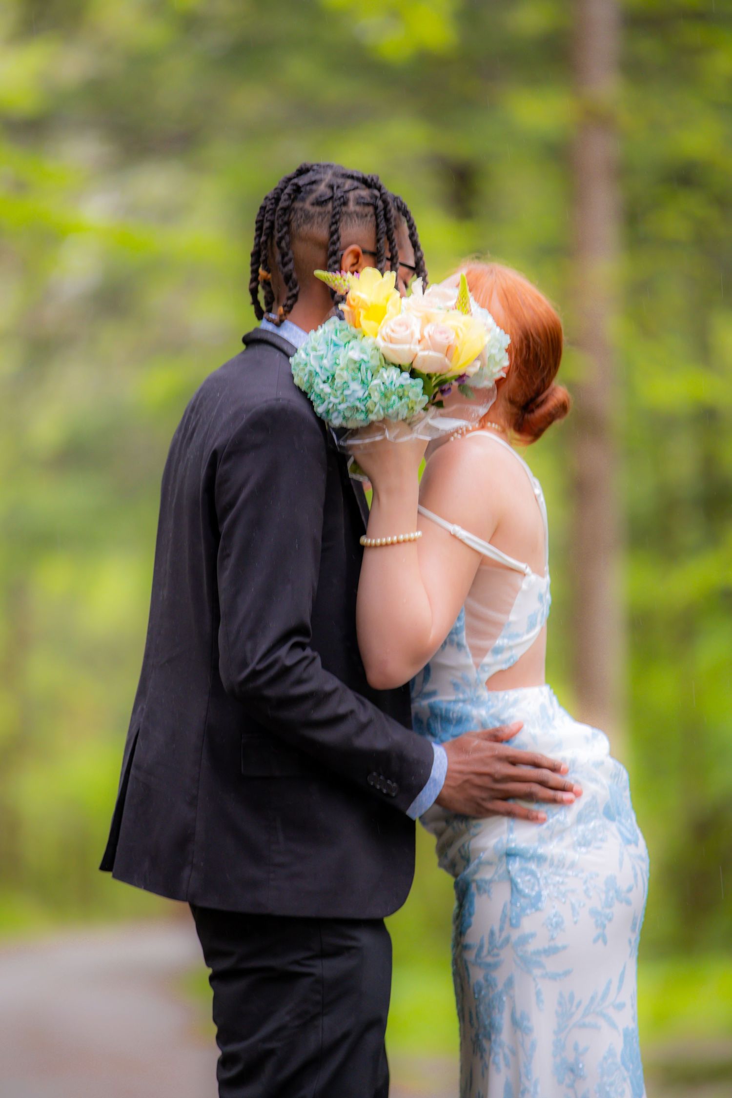 Couple in formal attire kissing behind a yellow and blue bouquet in a lush green forest setting.