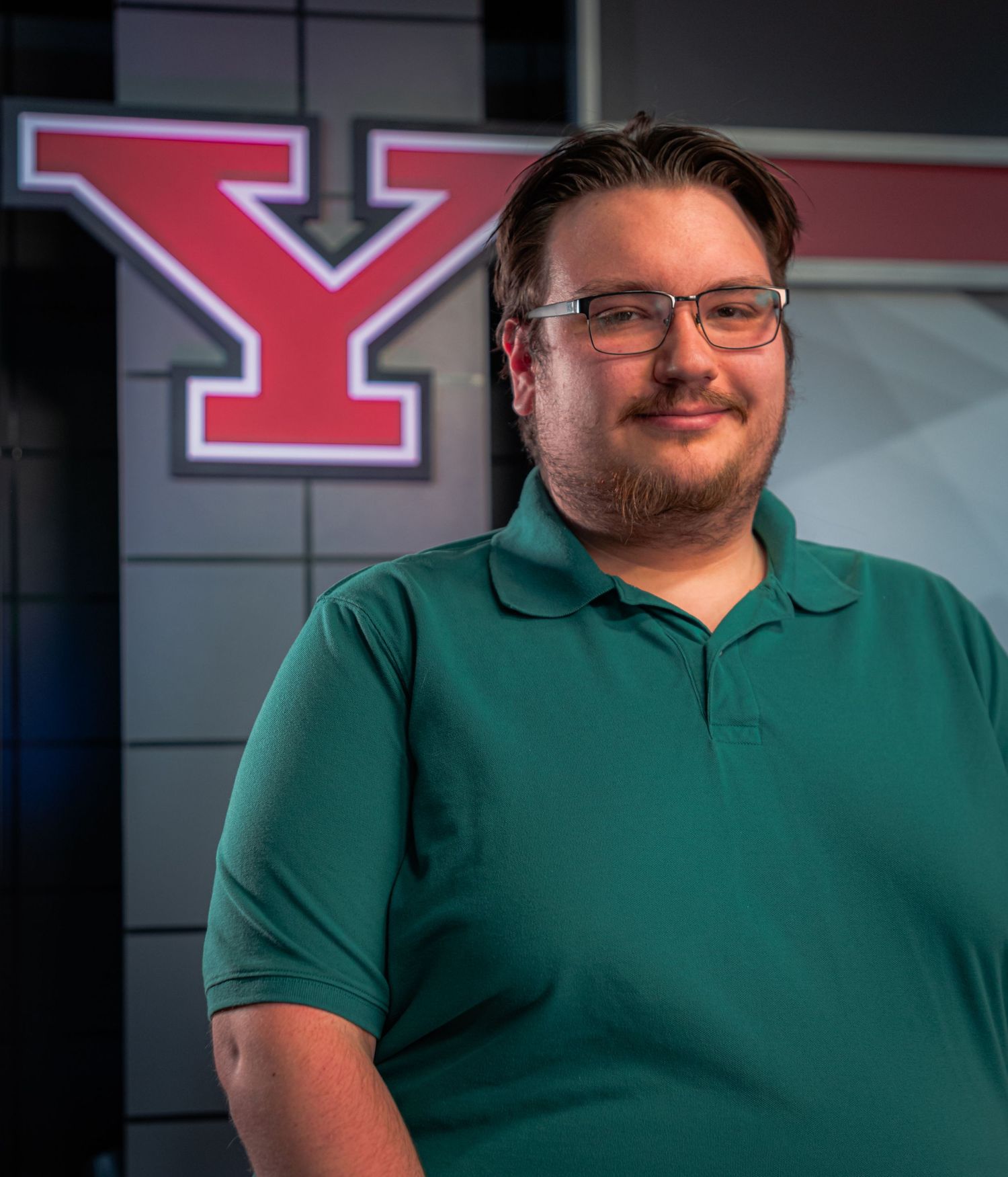 A young man wearing glasses and a green polo shirt smiles in front of a Youngstown State University Y logo.