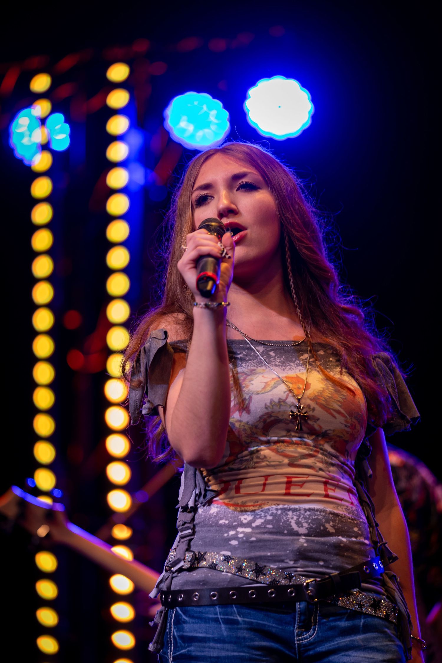 Young female singer performing on stage with microphone, colorful stage lights glowing behind her.