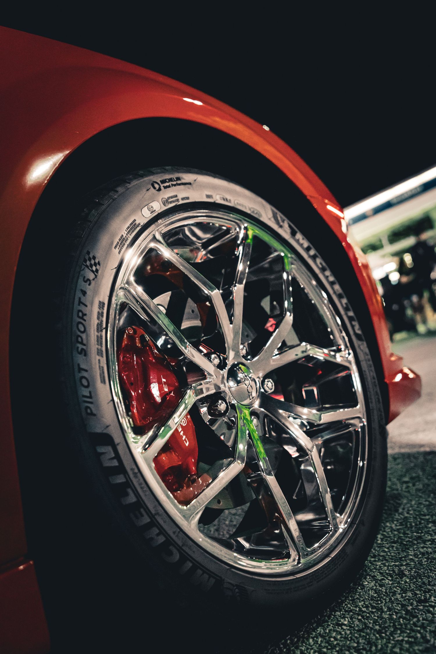 Close-up of a chrome sports car wheel with Michelin Pilot Sport tire and red brake calipers at night.