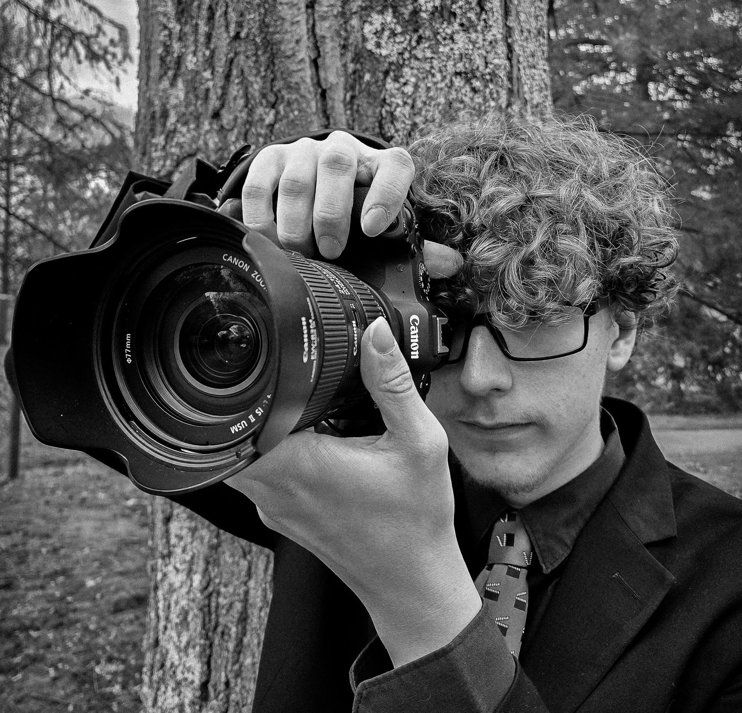 Black and white photo of a photographer holding a DSLR camera against a natural outdoor background.