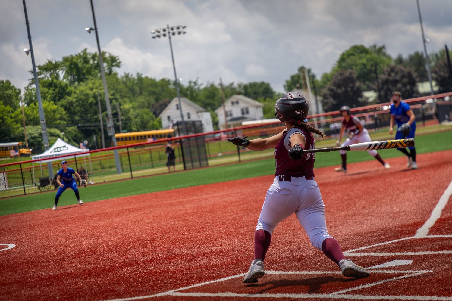 Softball players compete on a red turf field during a game under cloudy skies.