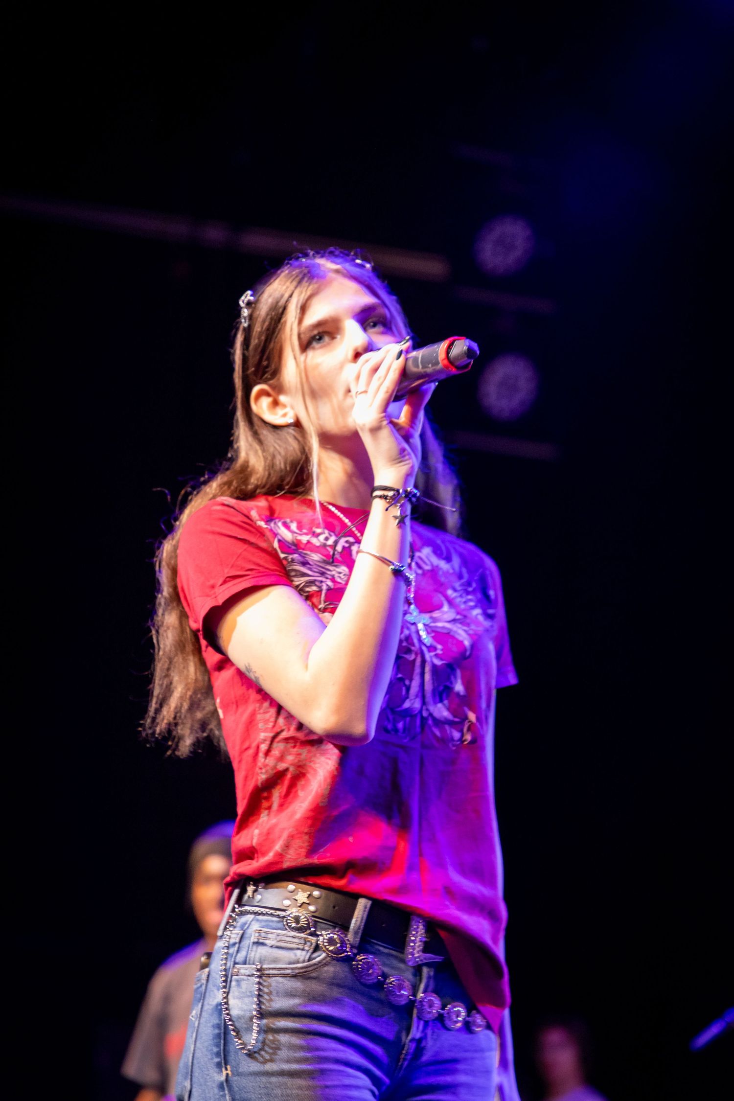 Young woman singer performing on stage holding microphone wearing red t-shirt and jeans under colorful concert lights.