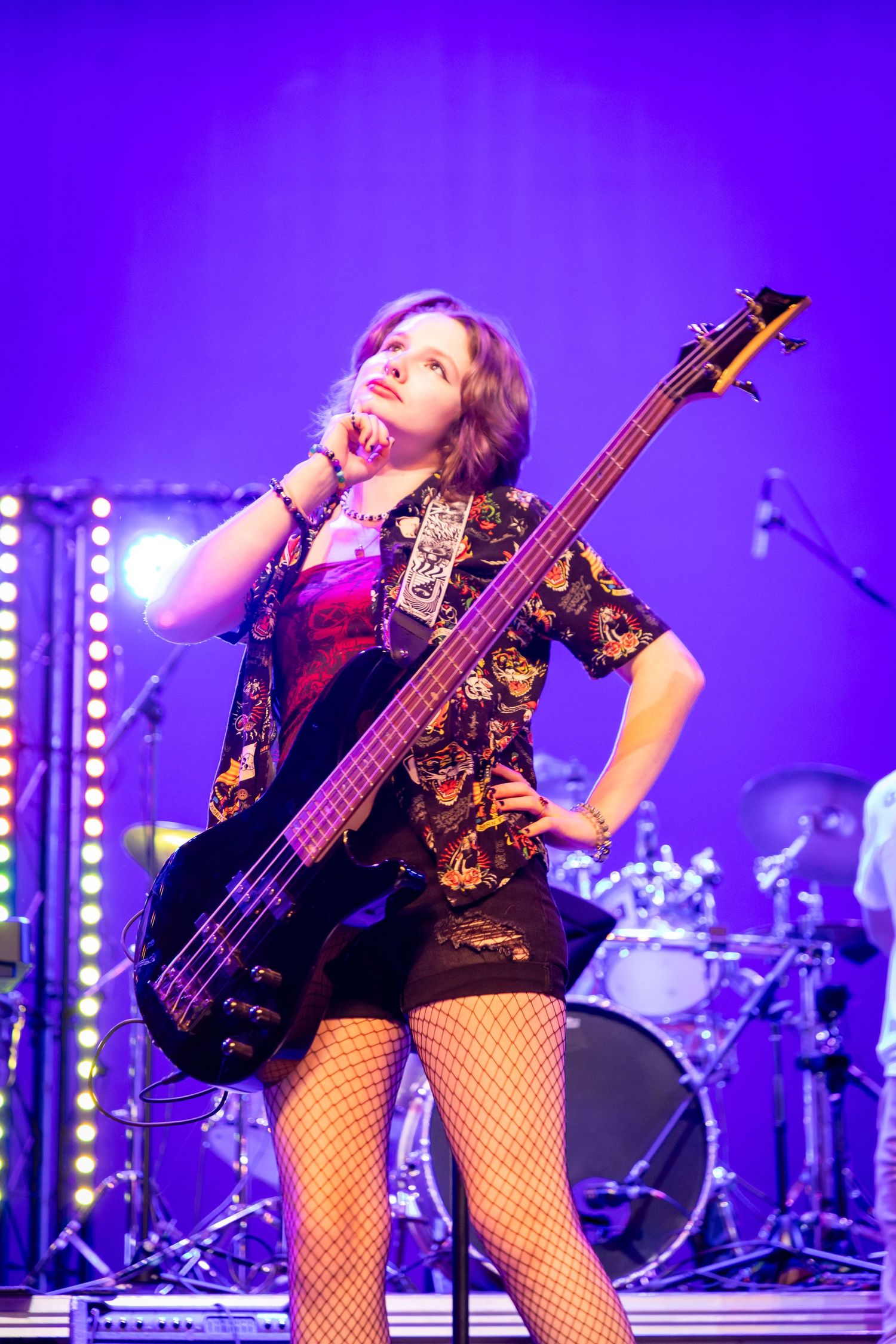Young woman bassist in fishnet stockings poses confidently on stage with electric bass guitar under purple lights.