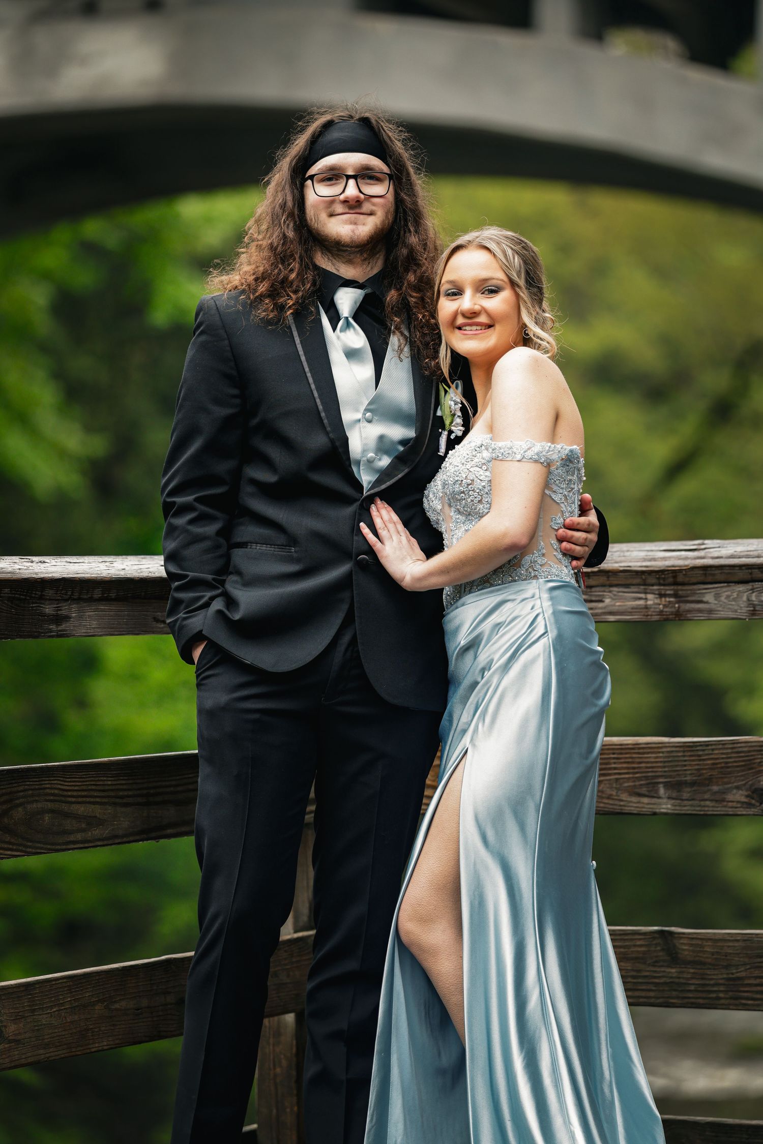 A couple in formal prom attire poses on a wooden bridge, he in a black tuxedo and she in a silver-blue gown.