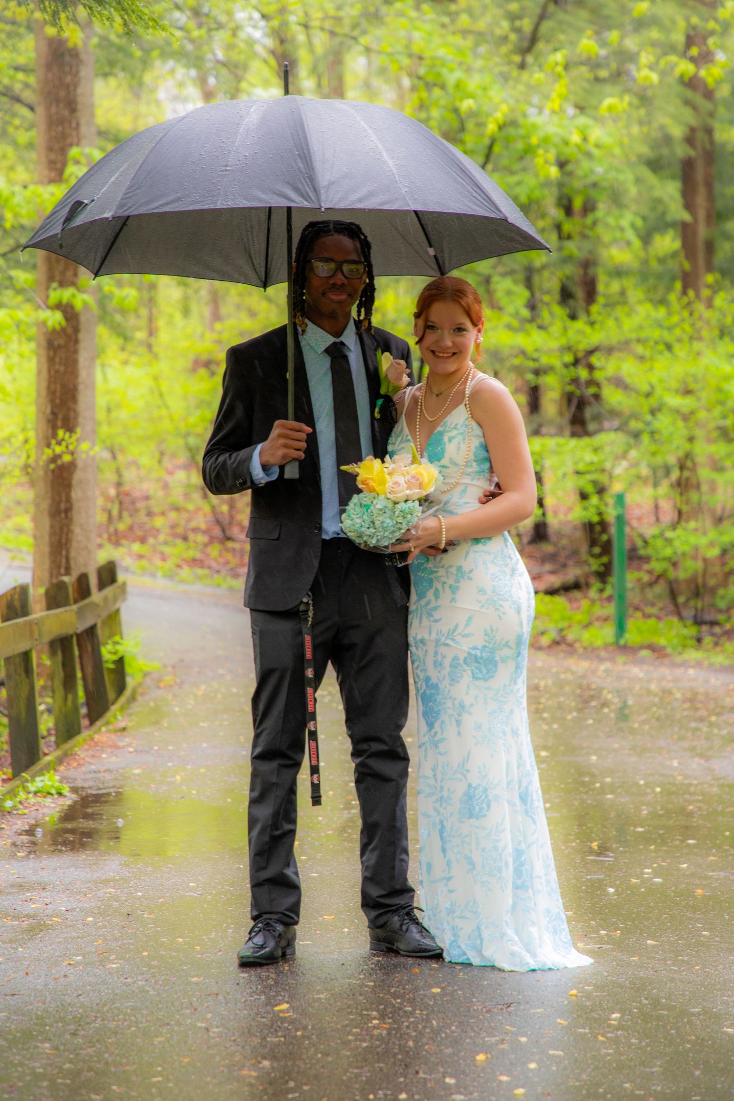 Couple in formal attire sharing umbrella in rain, woman in blue gown holding bouquet, man in black suit with boutonniere.