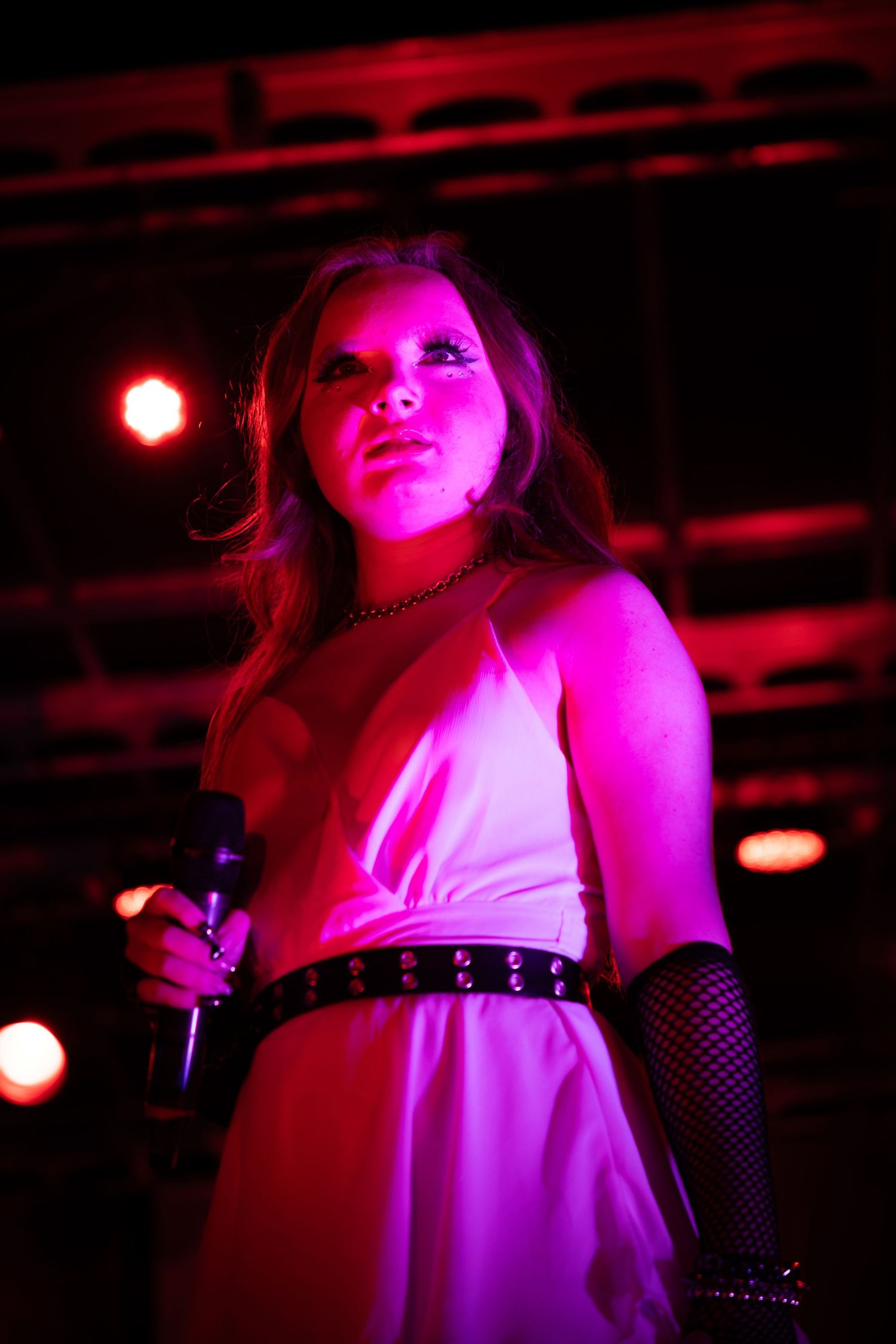 A female singer performs on stage under pink and red lights, wearing a white dress and fishnet gloves while holding a microphone.