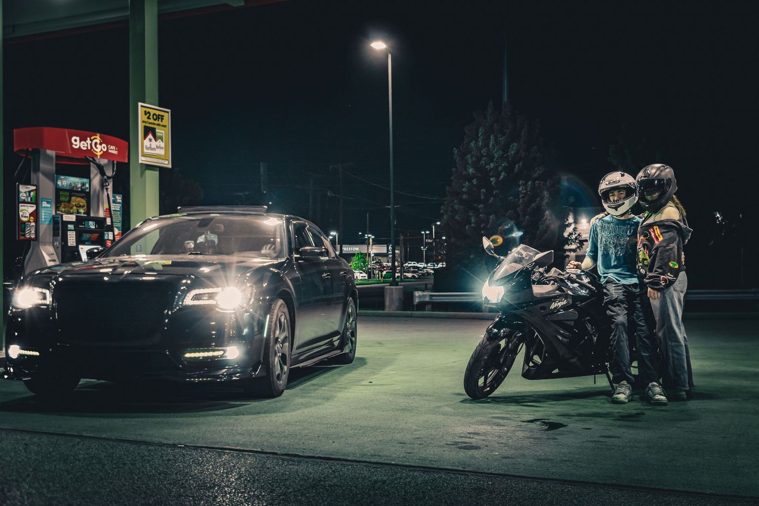Motorcyclists and a black luxury car meet at a gas station at night under bright lights.