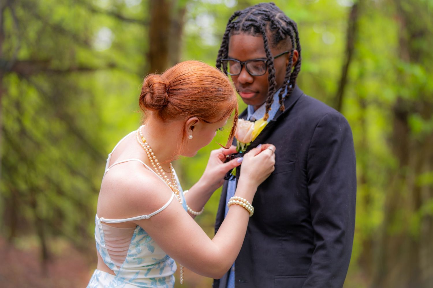 A young woman in a blue dress pins a boutonniere on a man in a suit outdoors before prom.