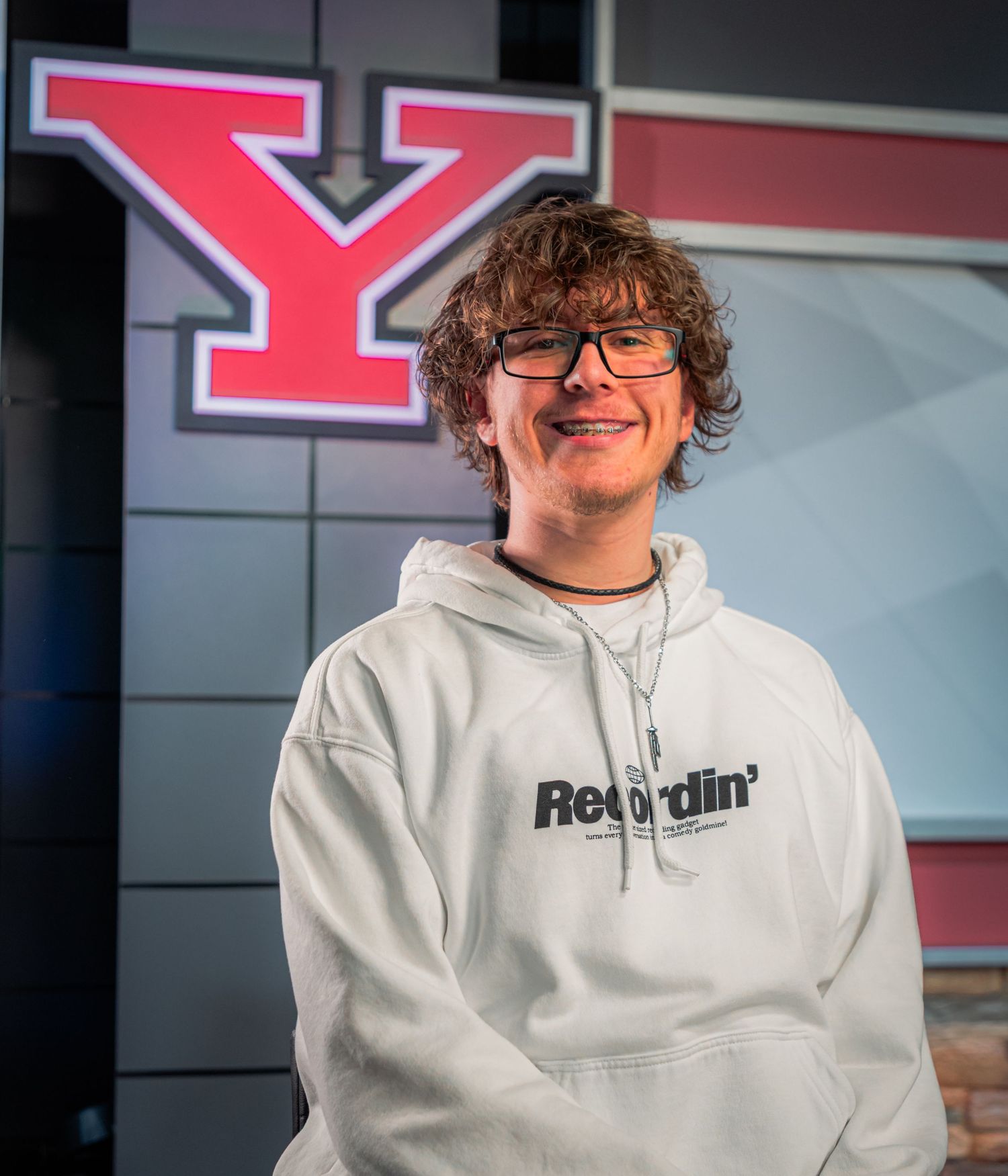 Young man in white Recordin hoodie smiling in front of a large red Y logo backdrop.