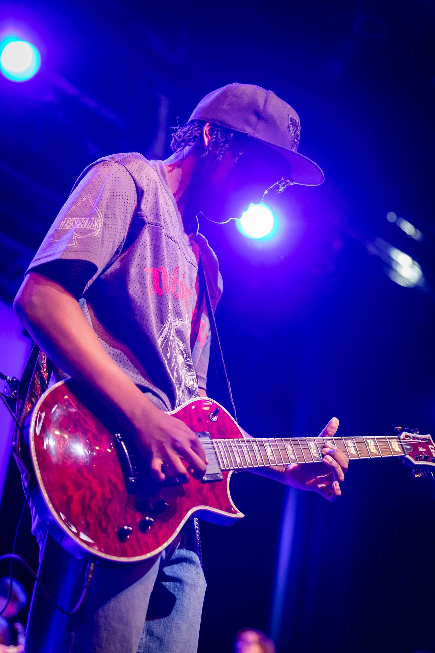 Guitarist playing red electric guitar on stage under blue concert lights wearing a gray cap and jersey.
