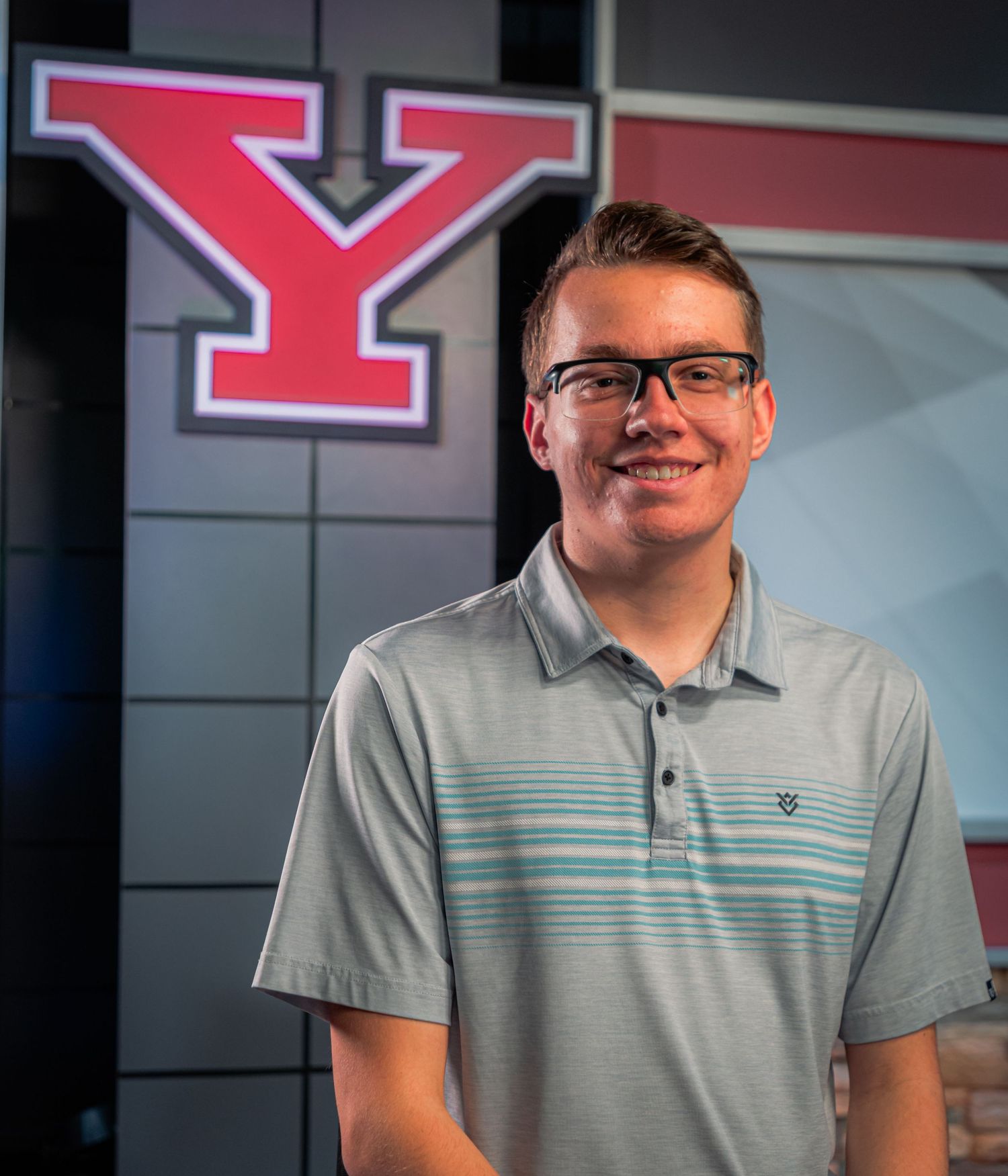 Young man in glasses smiling in front of a large red Y logo at Youngstown State University.