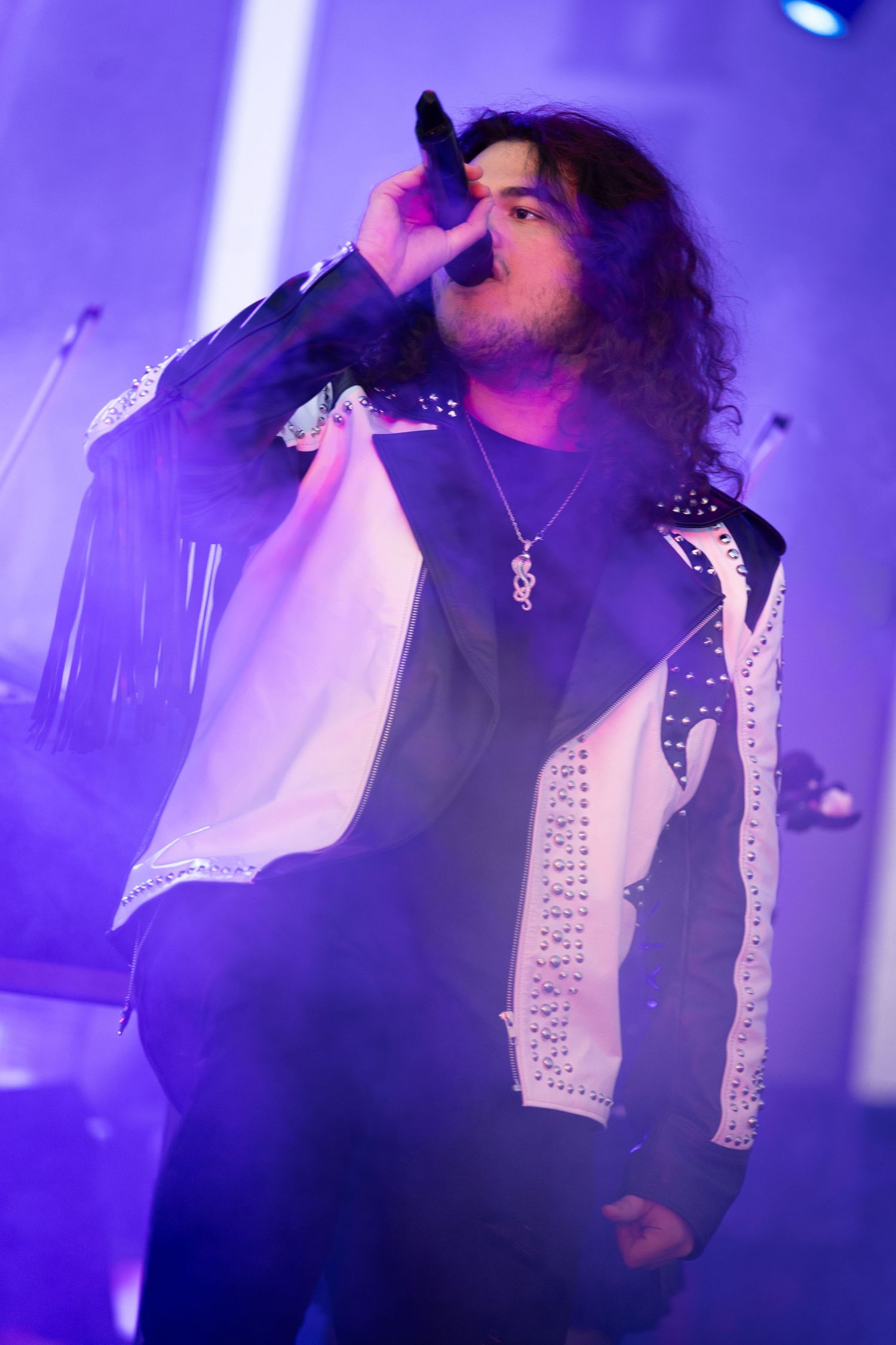 Male rock singer with curly hair performs on stage in a studded black and white jacket under purple stage lighting.