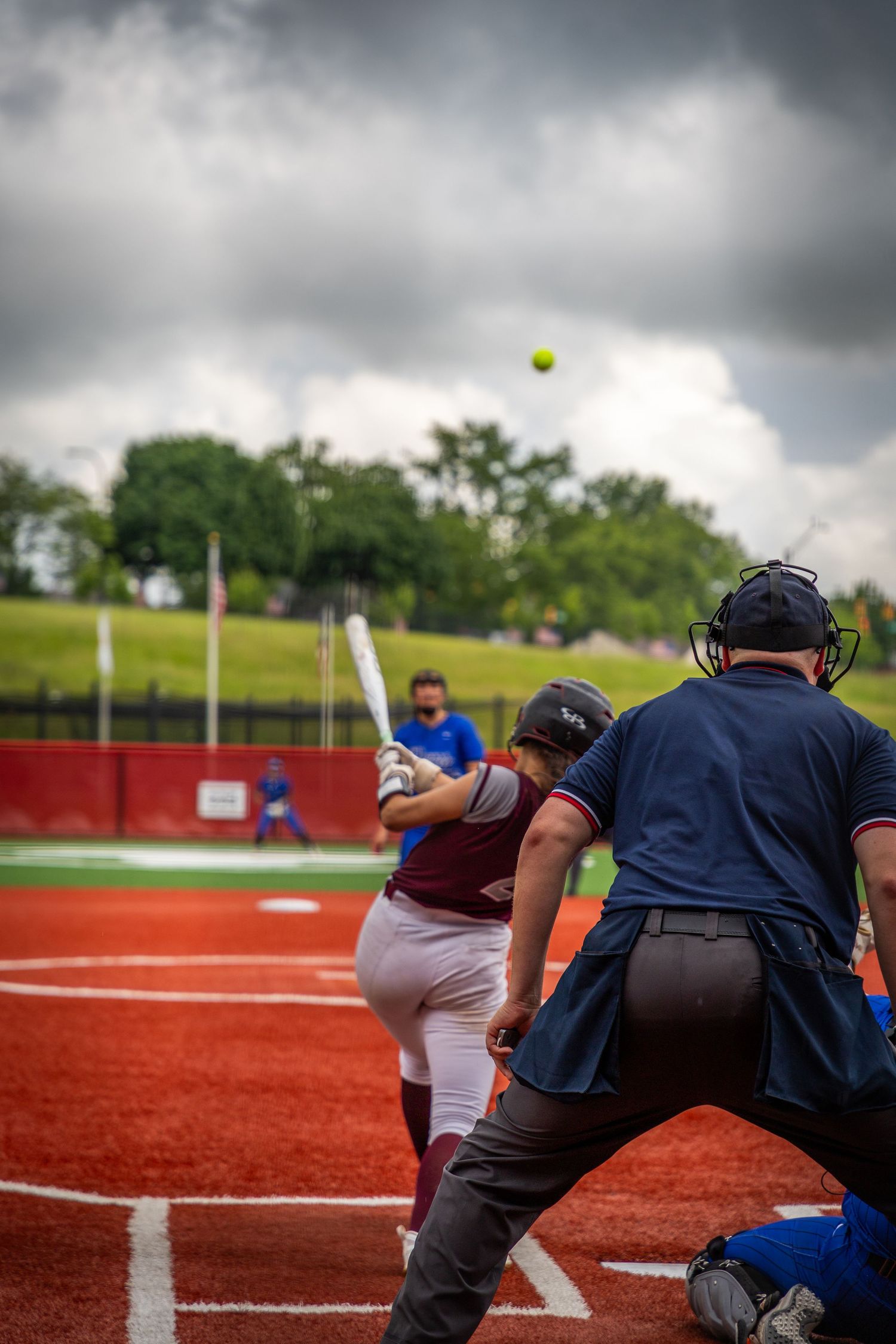 Softball game action shot with players and umpire on red turf field under stormy sky.