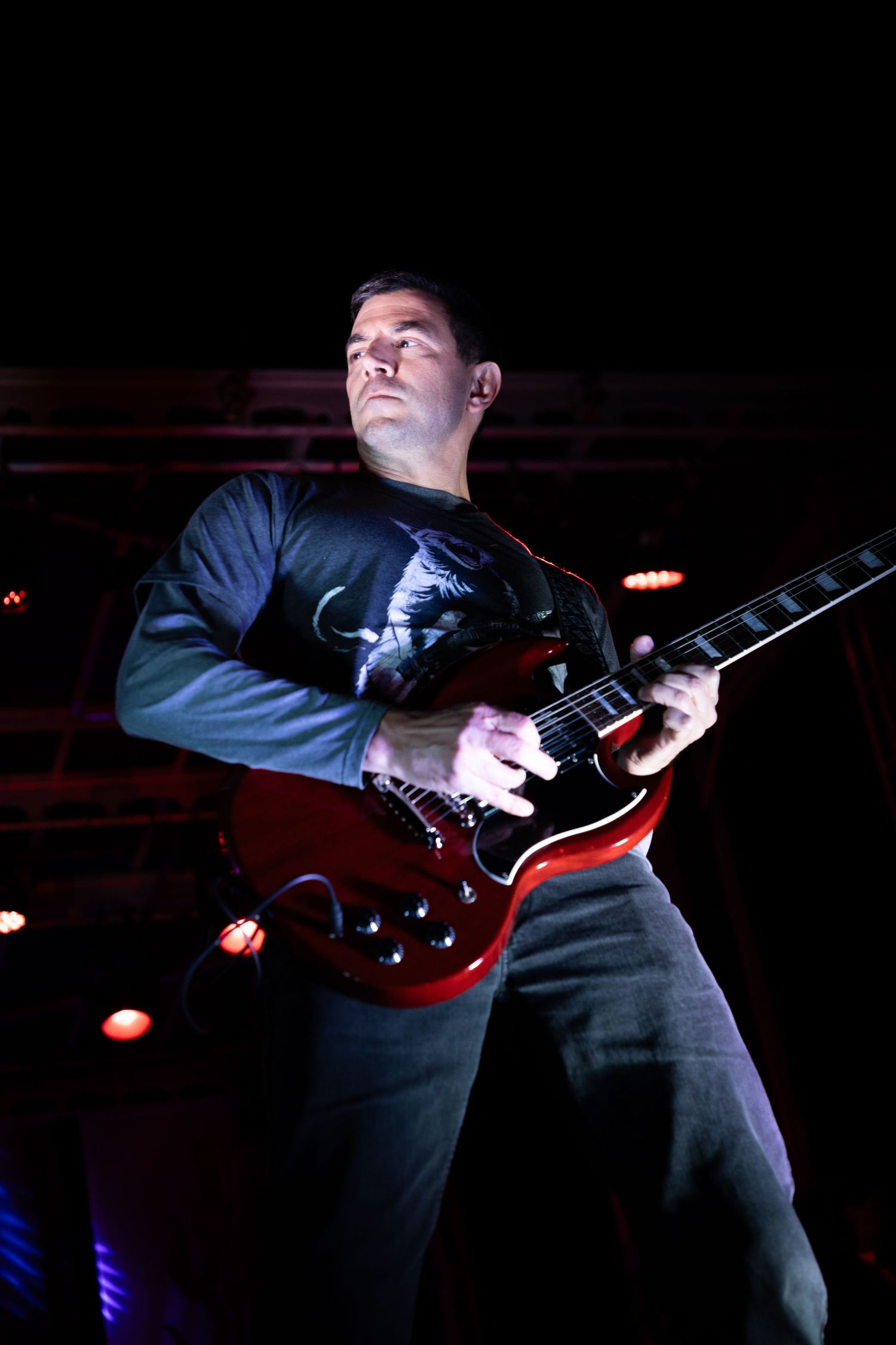 Male guitarist playing red electric guitar on dark concert stage under dramatic lighting.