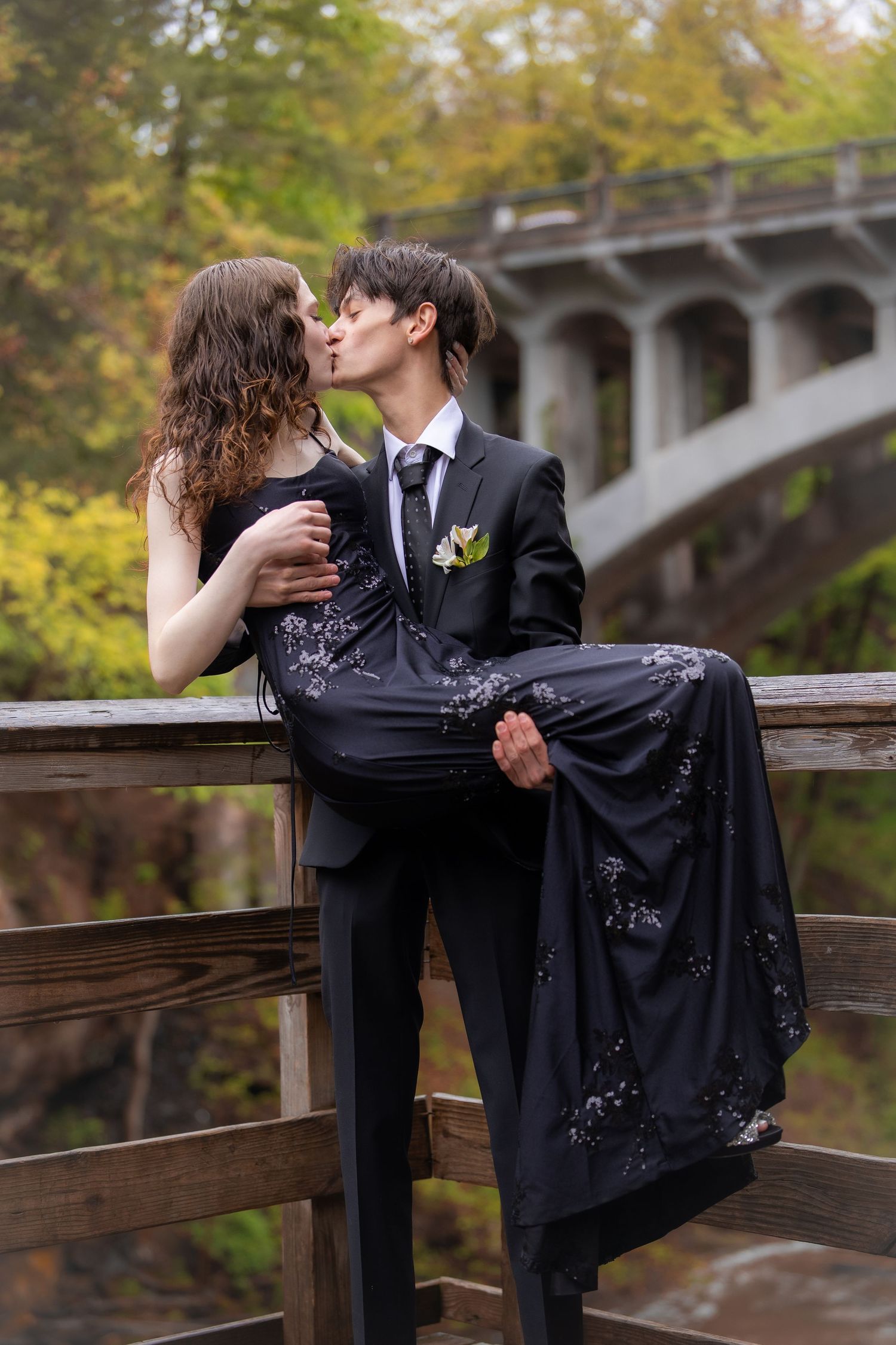 A couple in matching black formal attire share a kiss on a wooden bridge during prom photos.