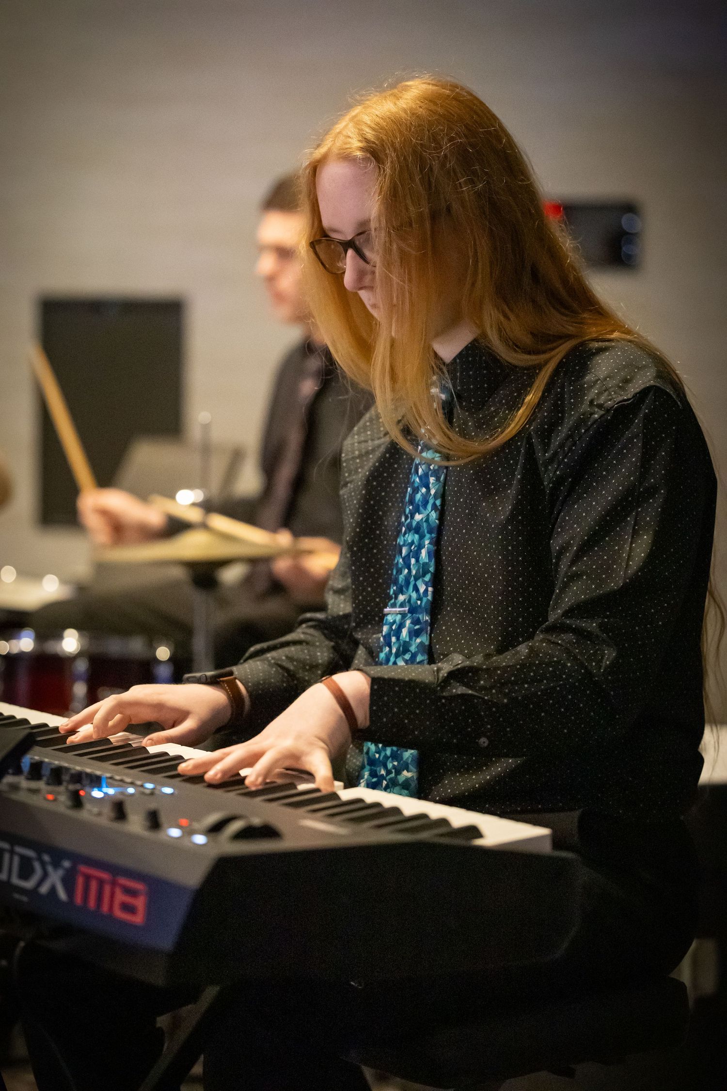 Red-haired musician playing a Roland keyboard synthesizer on stage with a drummer performing in the background.