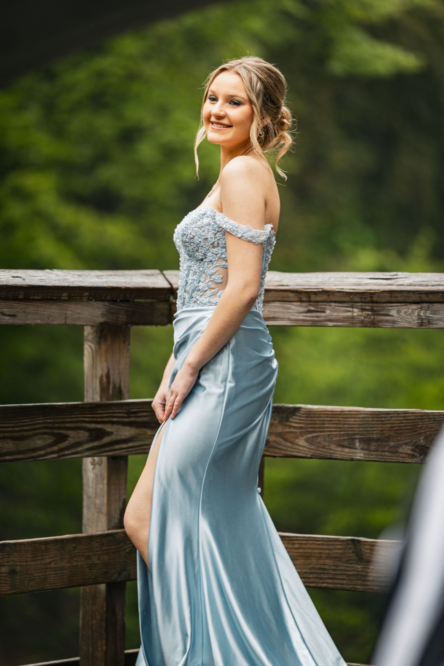 Young woman in light blue off-shoulder formal gown poses gracefully on wooden bridge surrounded by lush greenery.
