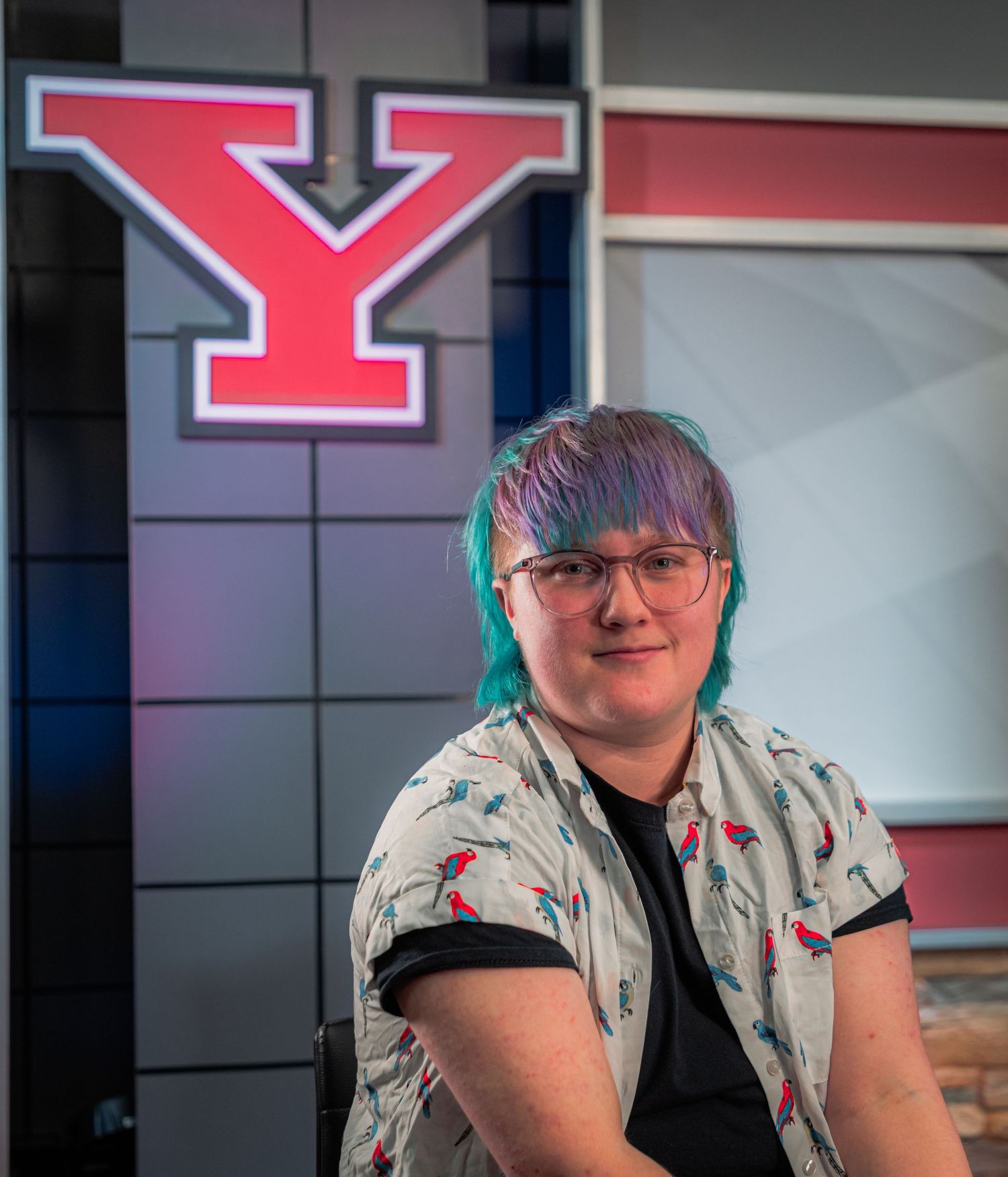 A person with colorful blue and purple hair and glasses smiles in front of a Youngstown State University Y logo.