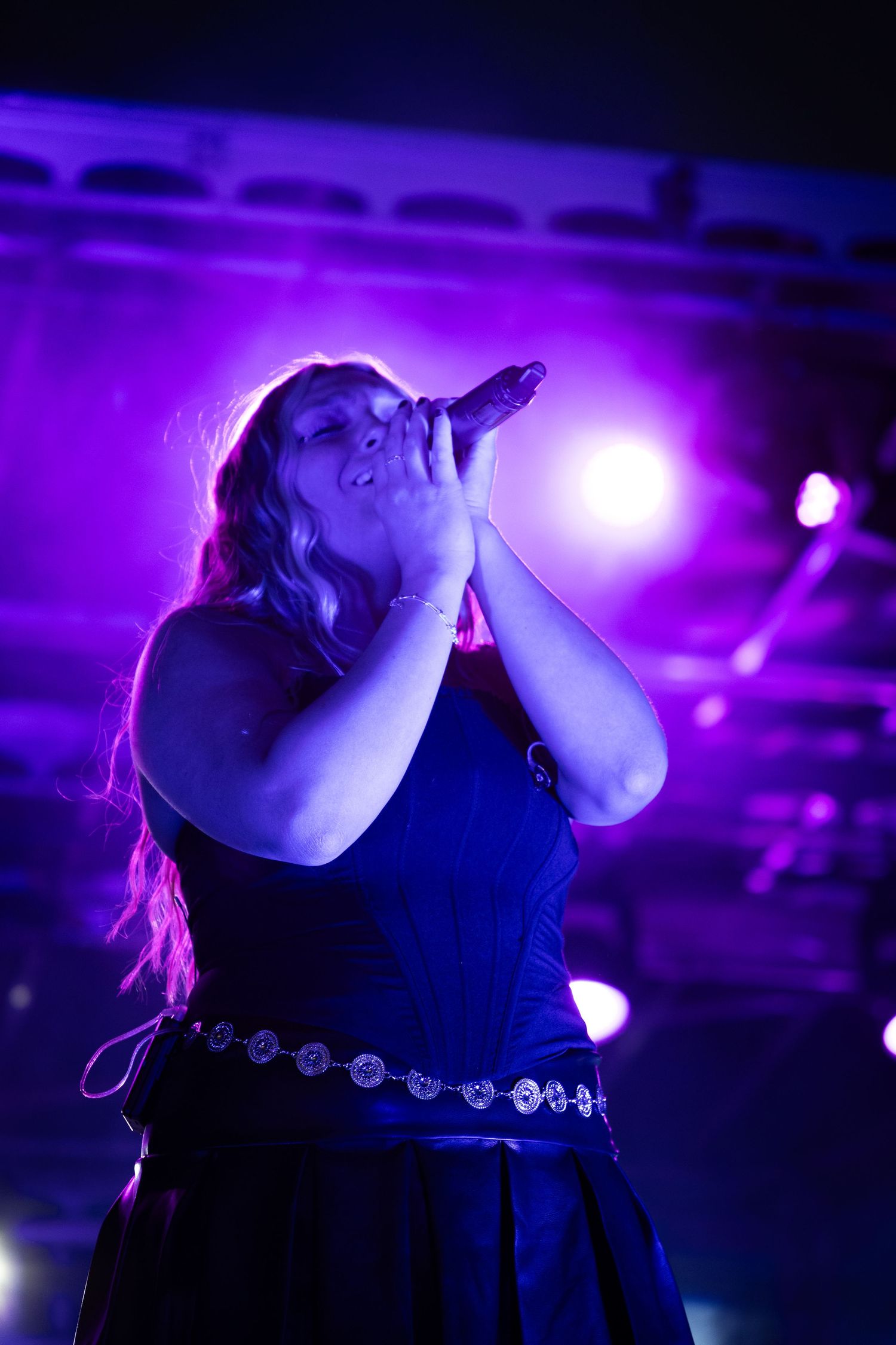 Female vocalist singing passionately into microphone under purple stage lights at a live music concert.