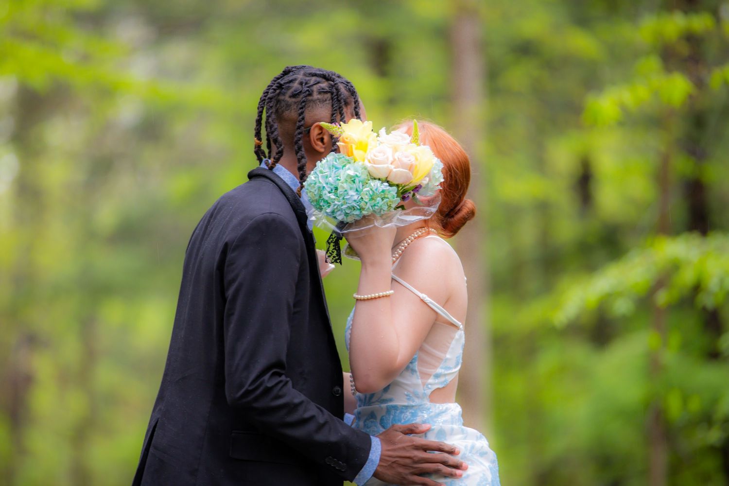 Couple sharing a kiss behind a colorful bouquet of yellow and blue flowers in a lush green forest setting.