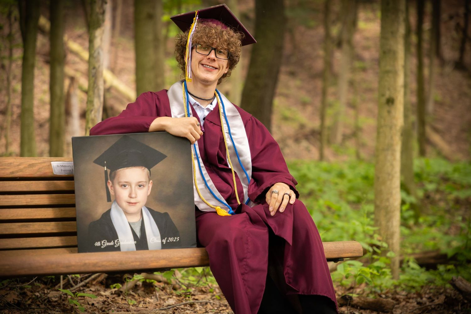 Graduate in burgundy cap and gown poses with portrait in wooded outdoor setting.